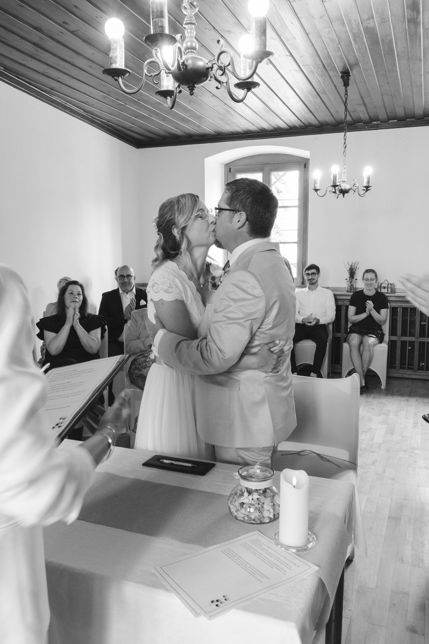 Black and white photo of bride and groom sharing first kiss at civil wedding ceremony before seated guests.