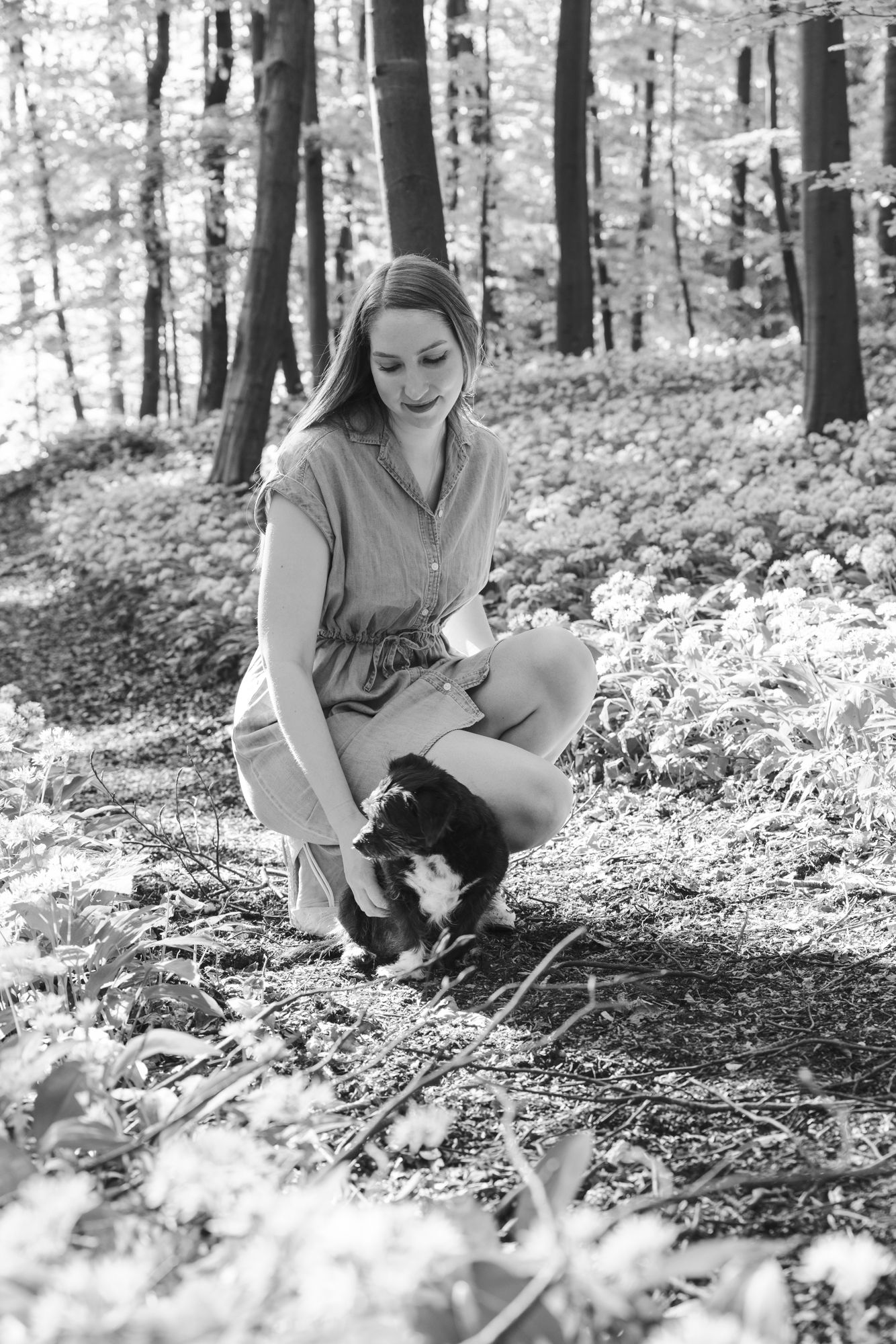 Black and white photo of a young woman crouching in a forest, gently petting a small cat among wildflowers.