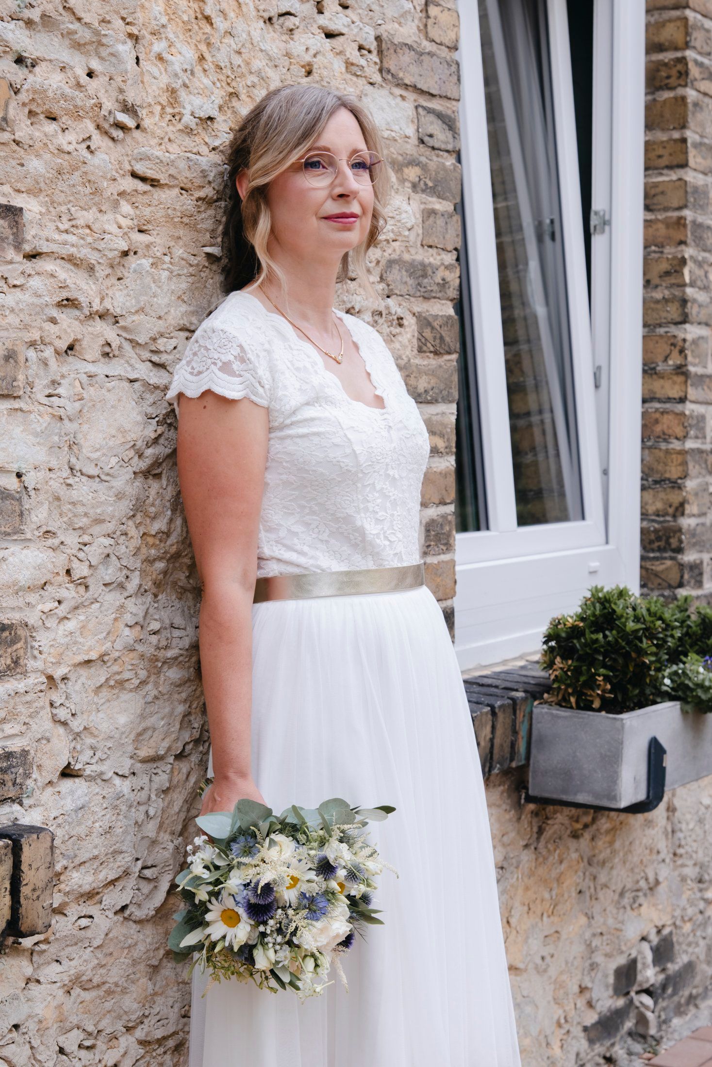 Elegant bride in white lace dress with gold sash holding bouquet, gazing upward beside stone wall and window.