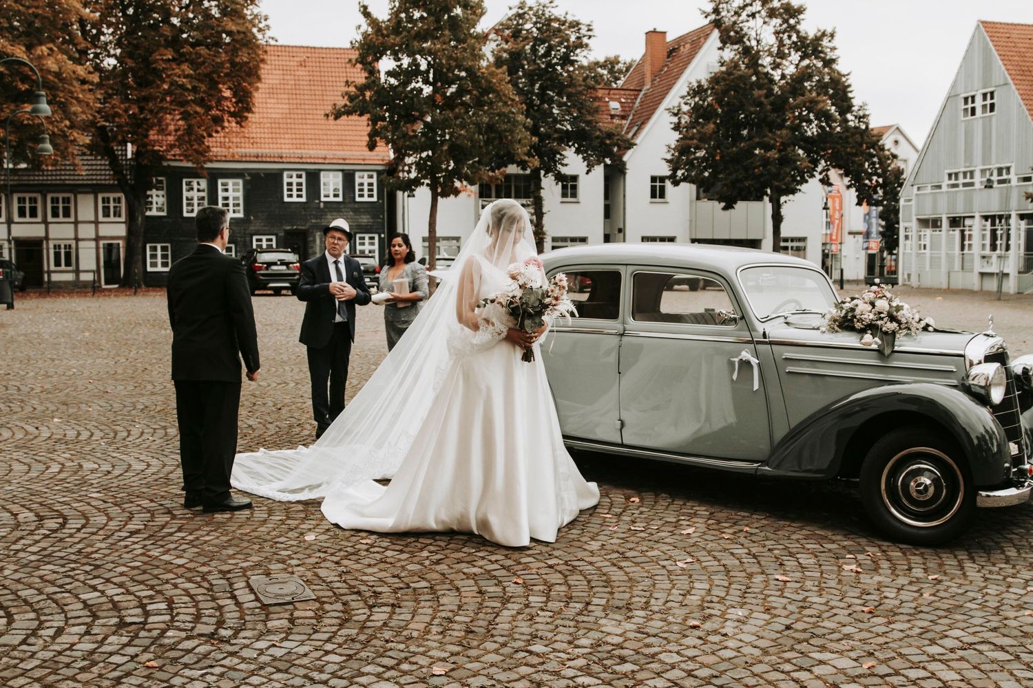 Bride in a flowing white gown arriving in a classic vintage car at a cobblestone European town square.