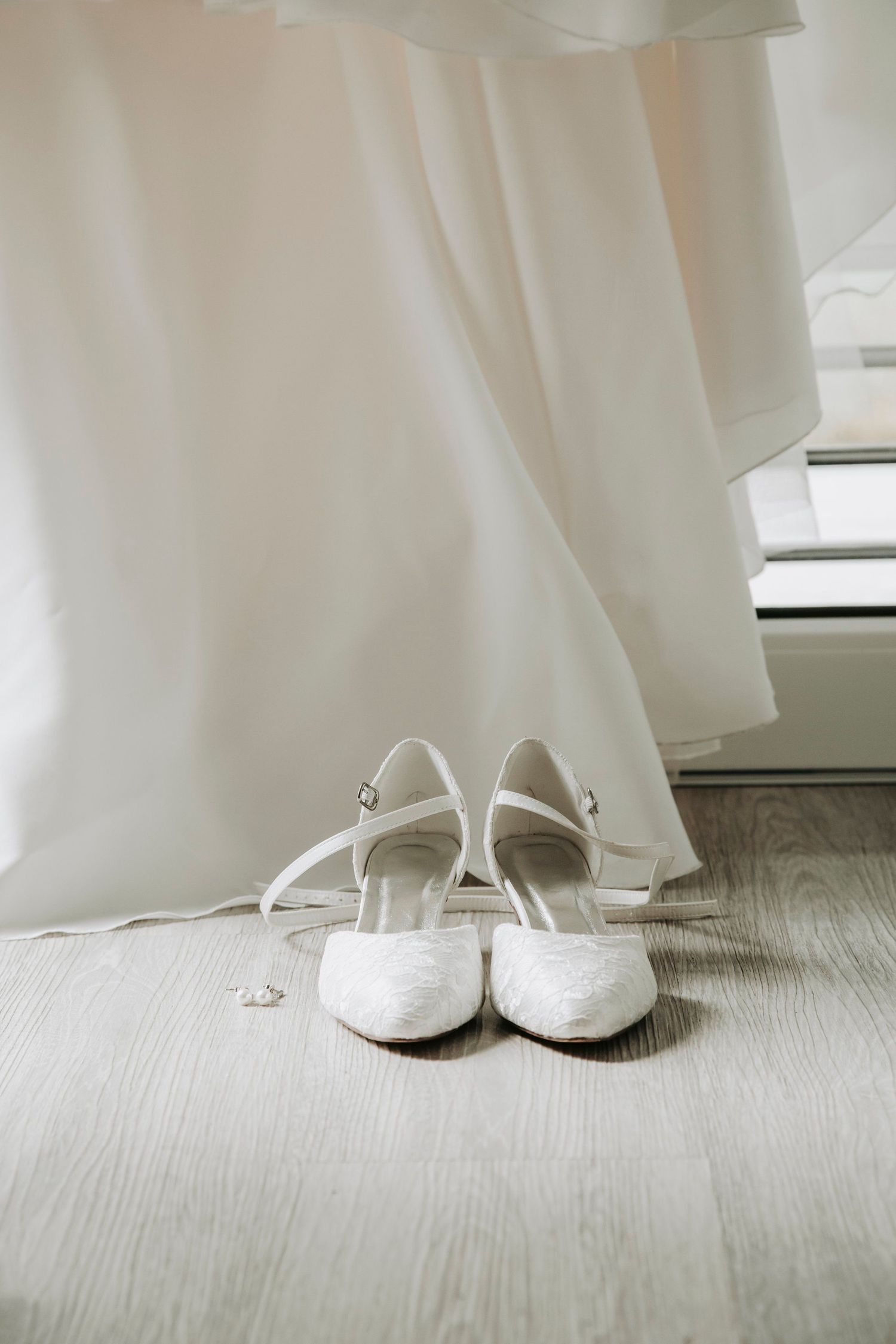 White lace bridal heels with ankle straps placed on floor beneath hanging wedding dress.