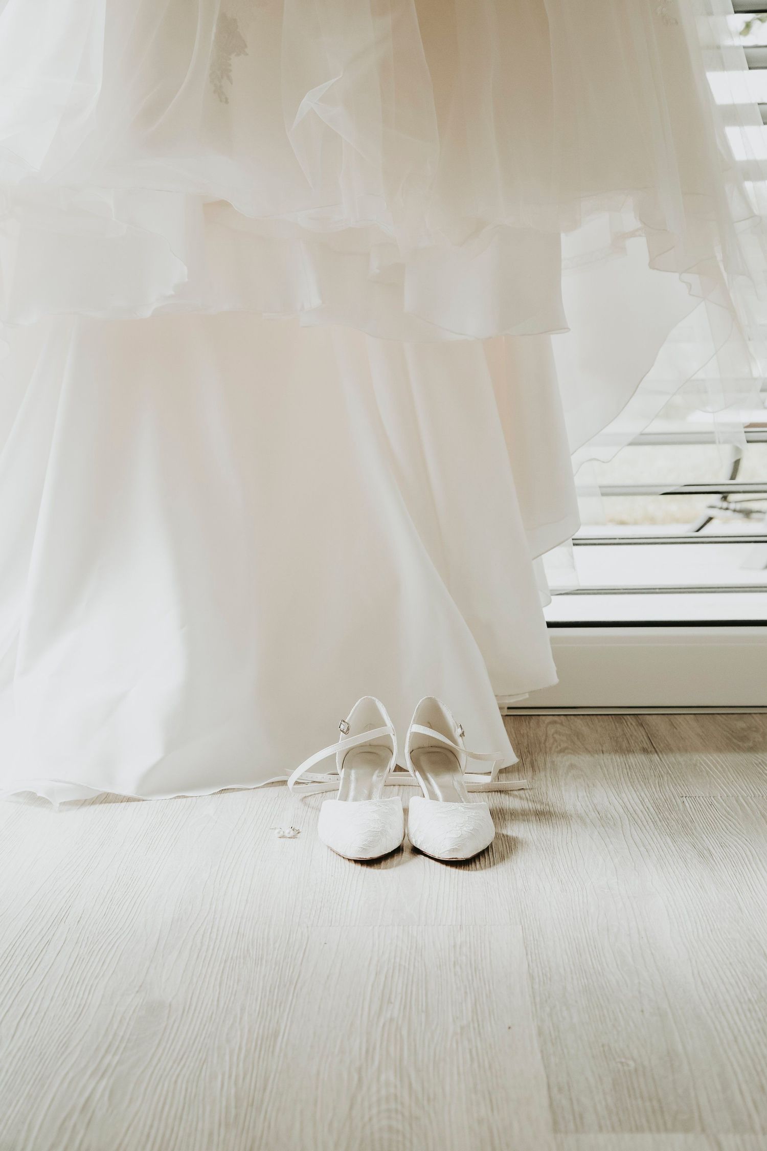 White bridal heels with ankle straps placed beside a flowing wedding dress near a bright window on a wooden floor.