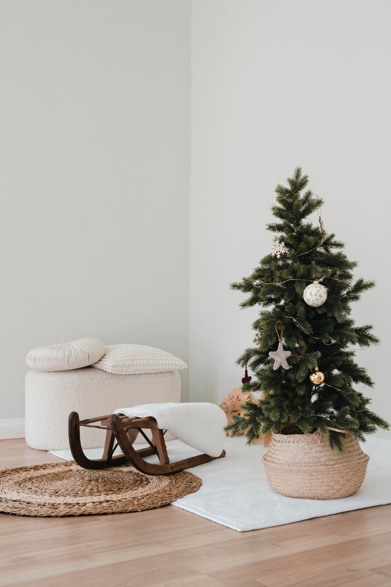 Minimalist Christmas room with decorated small tree in basket, vintage wooden sled on jute rug, and cream ottoman with pillows.