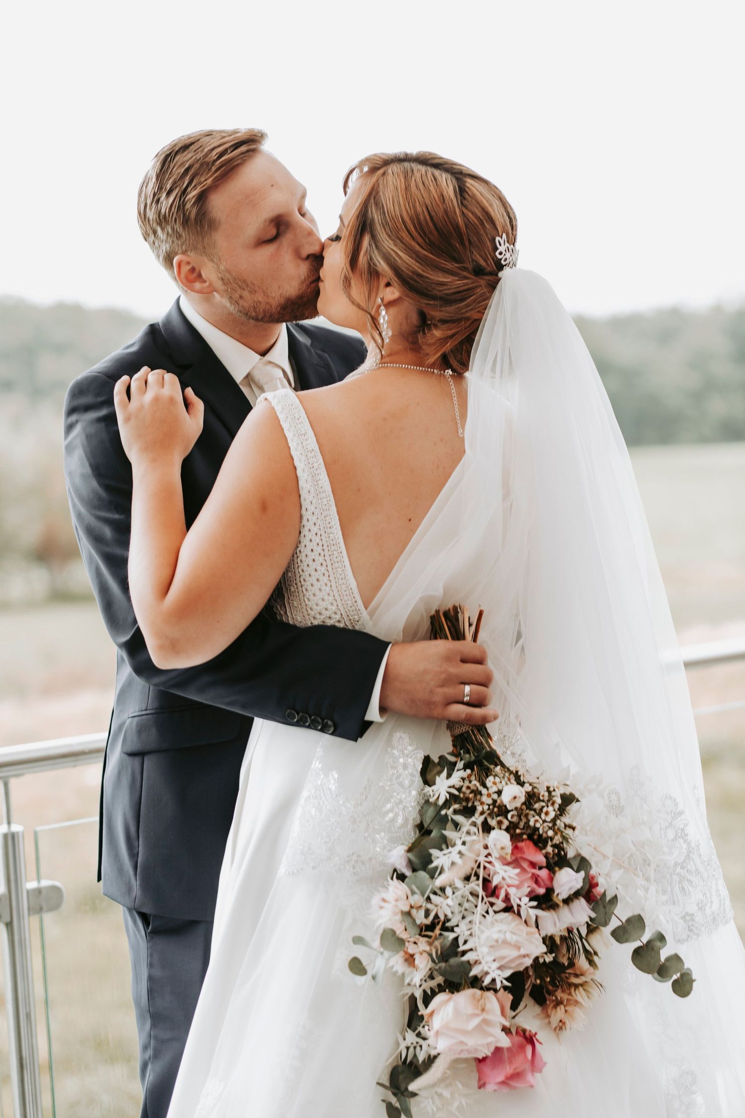 Bride and groom sharing a tender kiss on a balcony, bride holding a pink and white floral bouquet.