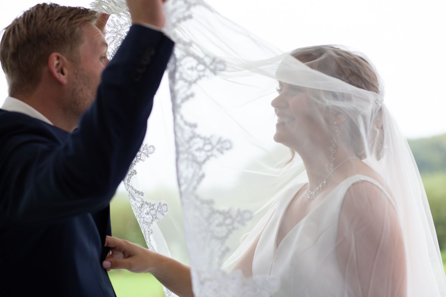 Groom lifting lace wedding veil over smiling bride in a tender romantic moment.