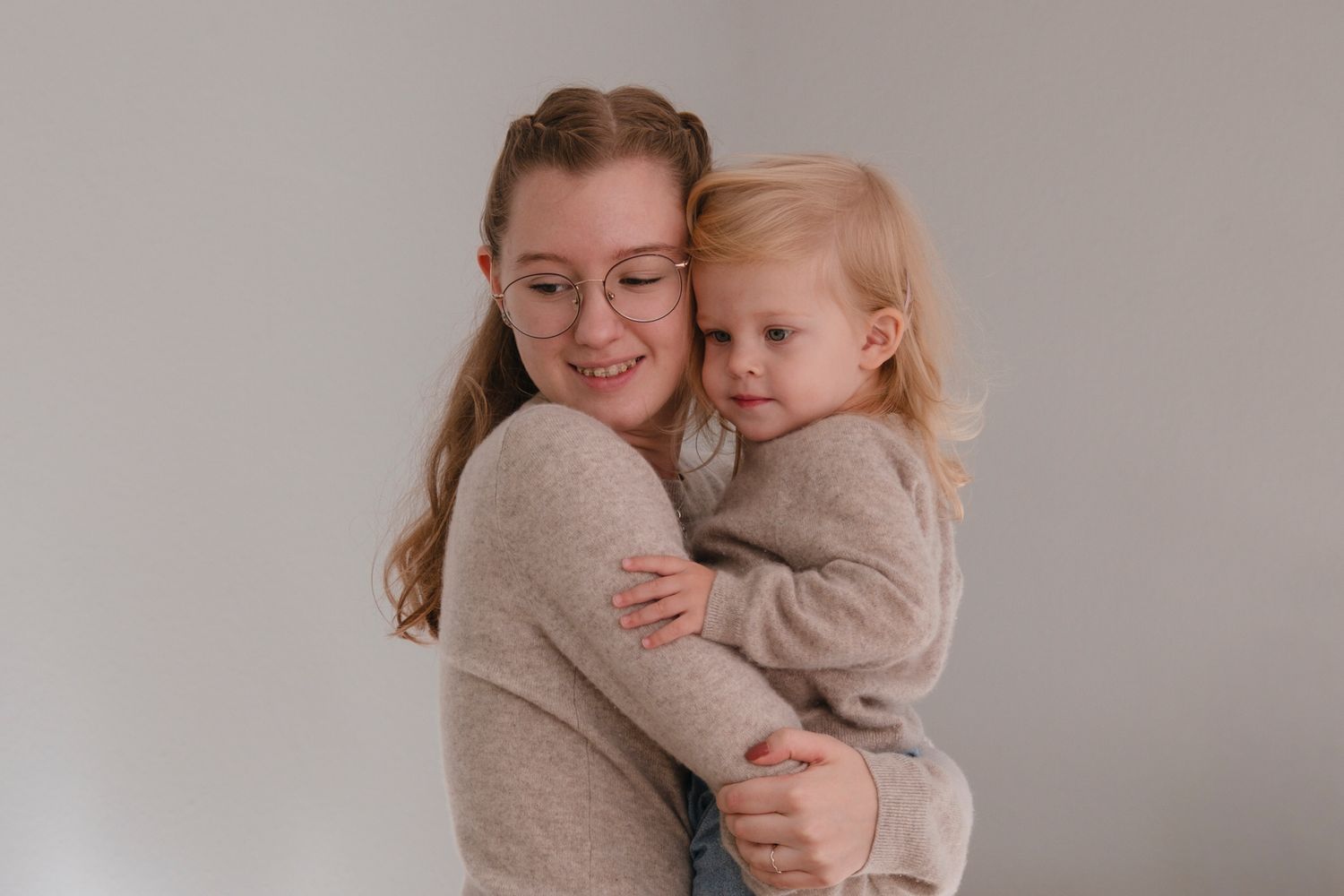 Smiling woman with glasses hugging blonde toddler girl against light gray background.