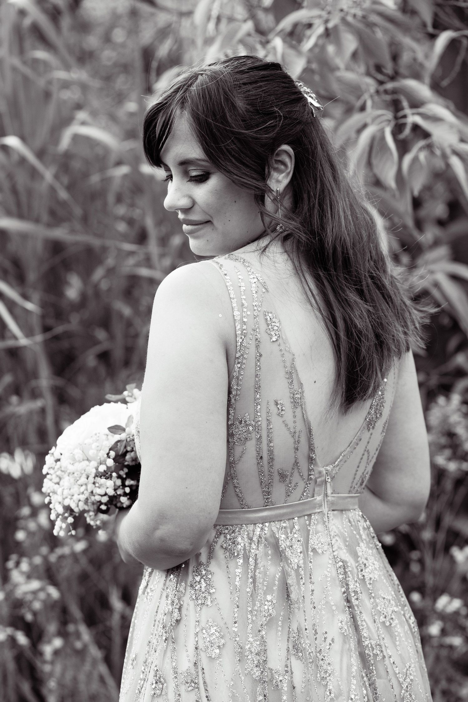 Bride in backless sequined gown holding bouquet, elegant black and white portrait in nature.