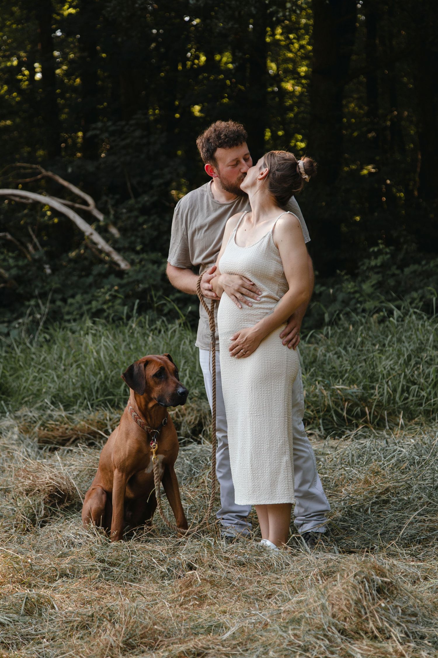 Couple kissing outdoors on hay with brown Rhodesian Ridgeback dog sitting beside them in forest.