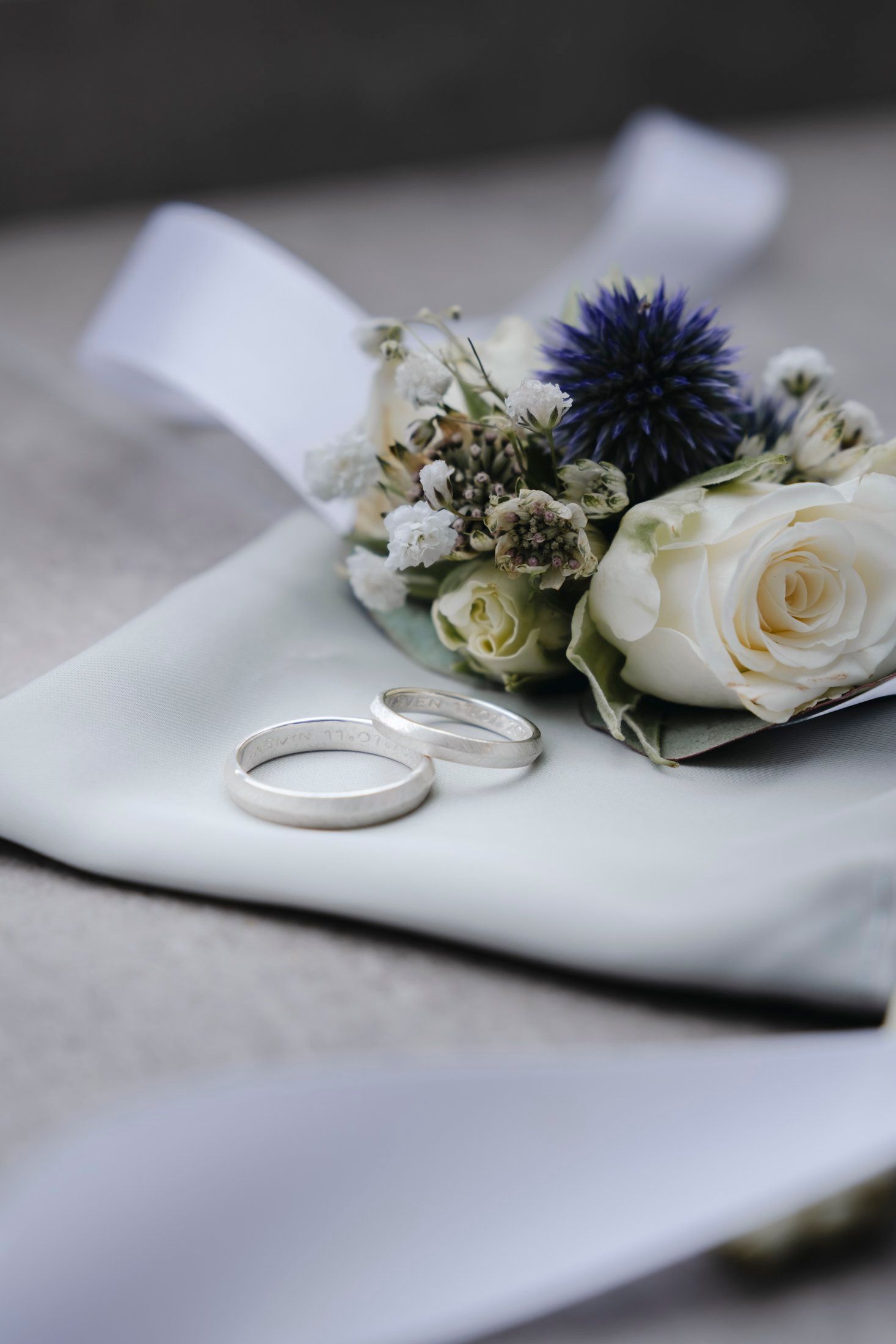 Two silver wedding rings placed beside a white rose and blue thistle boutonniere on fabric.