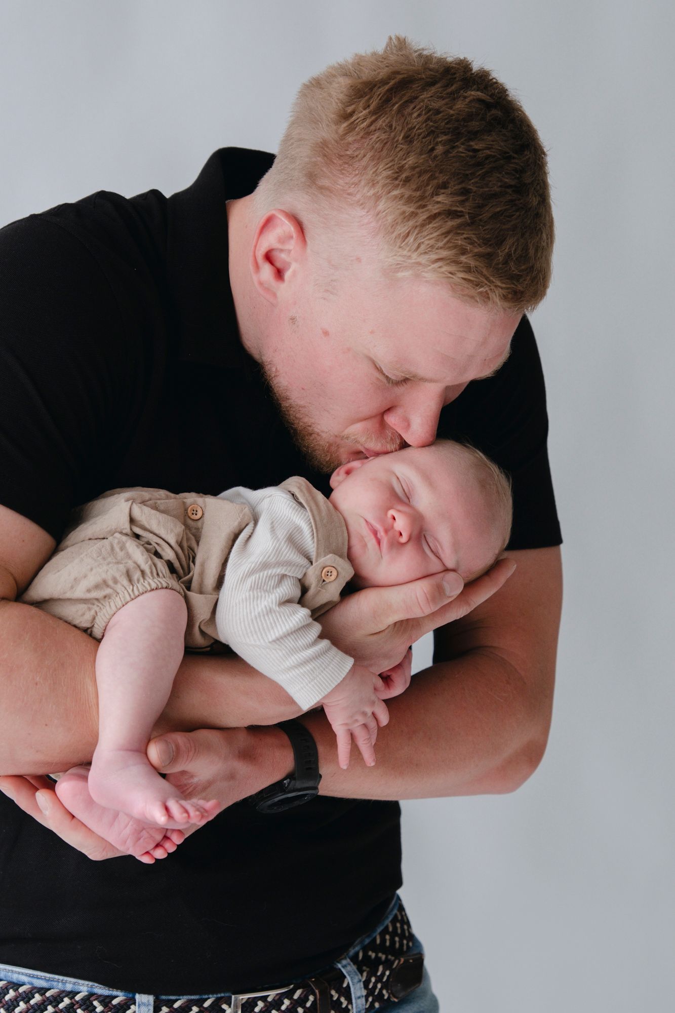 Father in black shirt tenderly kissing his sleeping newborn baby against white background.