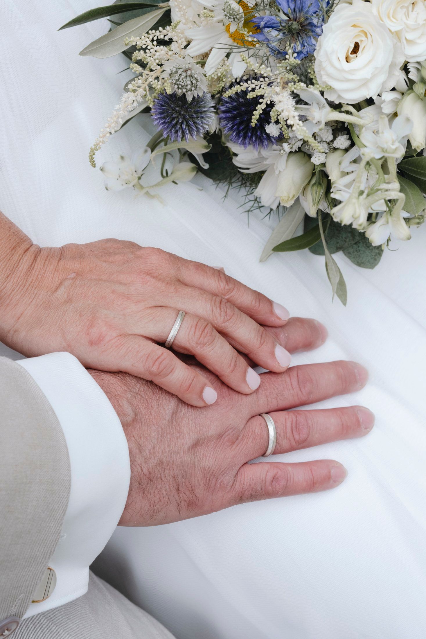 Close-up of newlyweds hands wearing matching silver wedding bands resting near white and blue bridal bouquet.