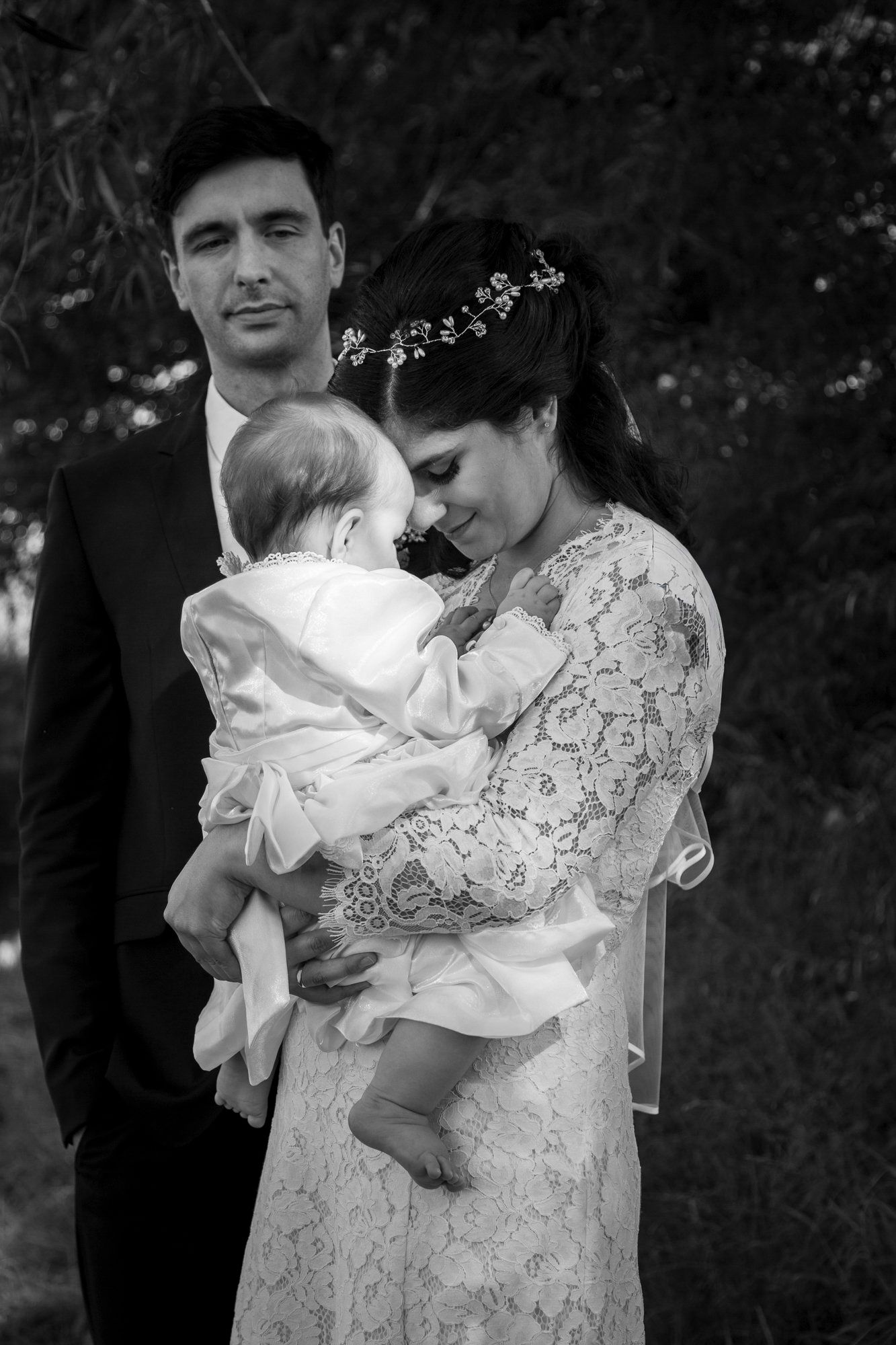 Bride and groom pose with their baby in a tender black and white family portrait outdoors after their wedding ceremony.