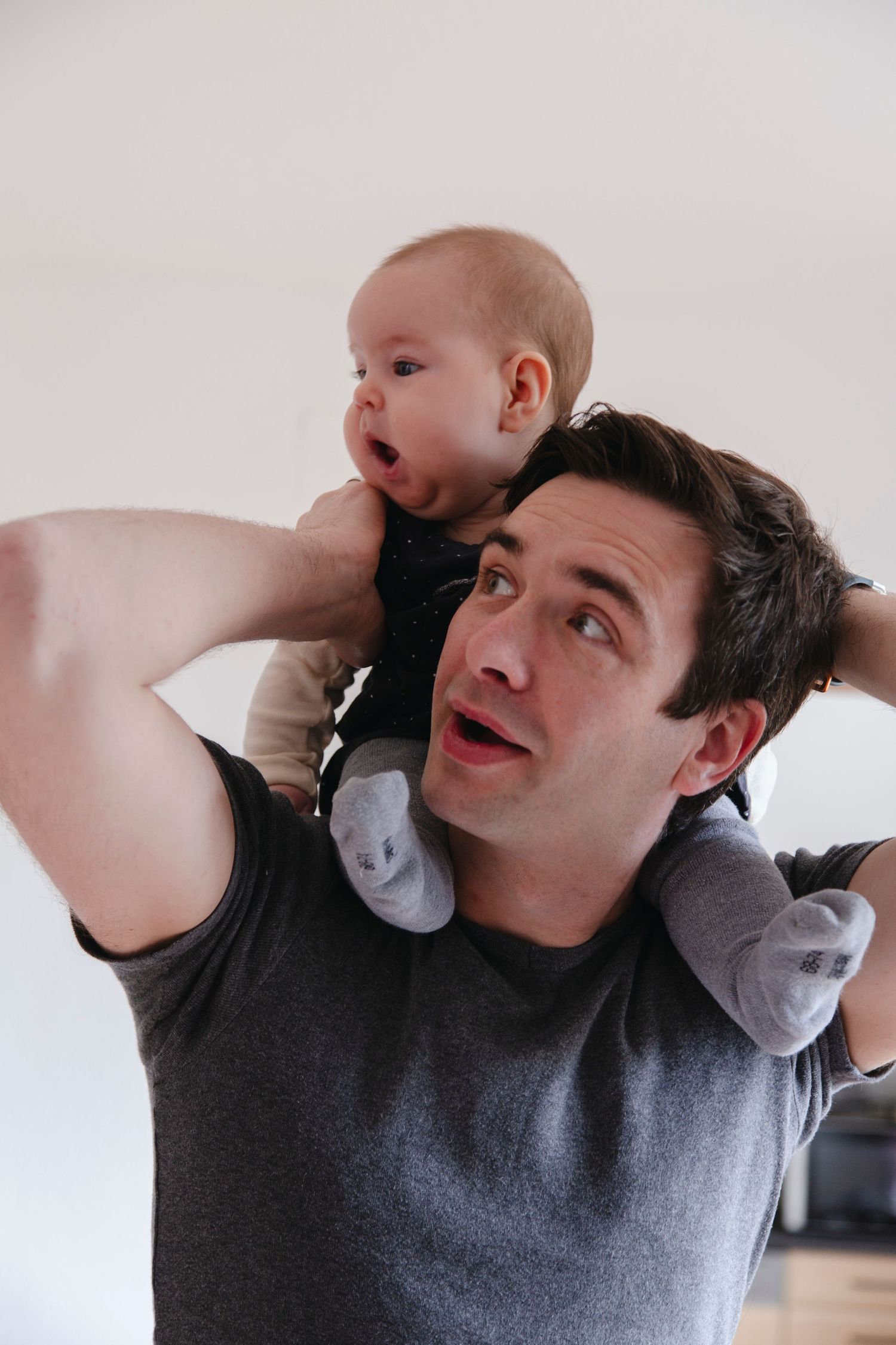 Playful father holding baby on his shoulders, both making surprised expressions against white wall.