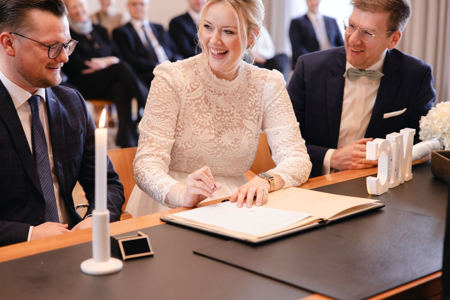 Smiling bride signs wedding register at ceremony table with groom and witness beside her.