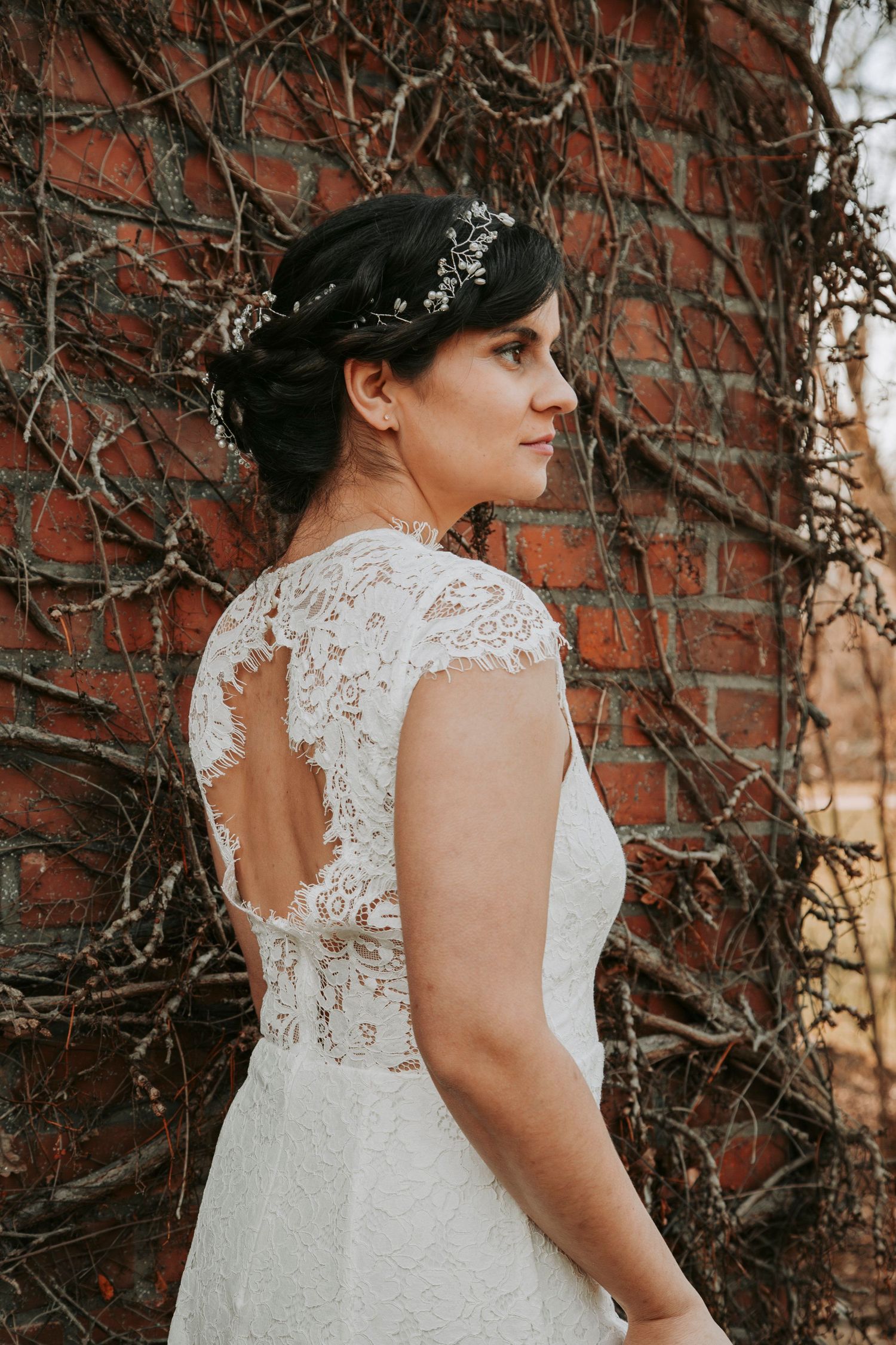 Bride in lace wedding dress with open back poses against brick wall covered in vines.