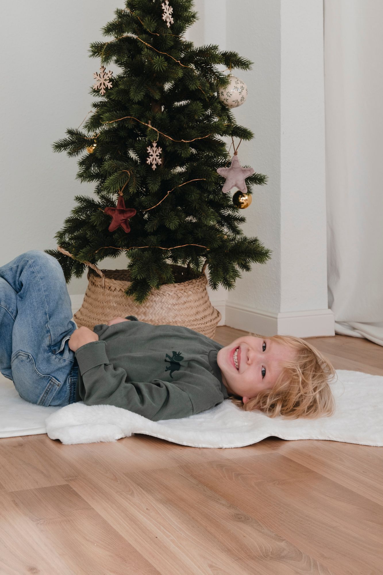 A smiling child with blonde hair lying on a white rug beside a decorated Christmas tree indoors.
