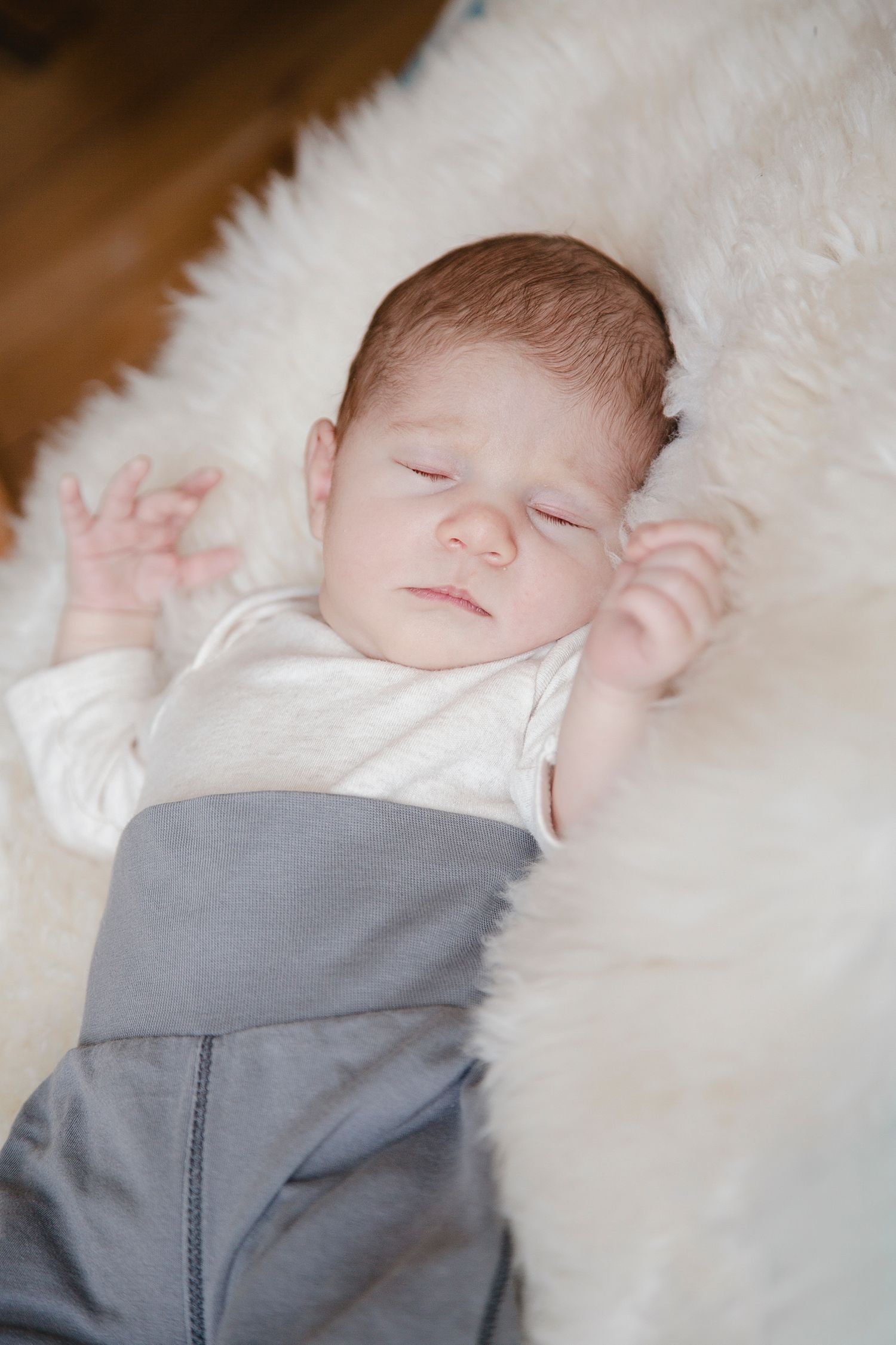 Sleeping newborn baby lying on a white fluffy fur rug in gray pants.