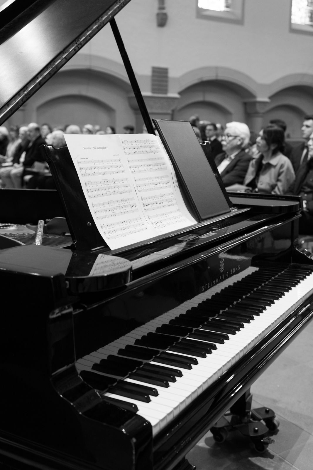 Black and white photo of grand piano with sheet music open before seated wedding ceremony guests.