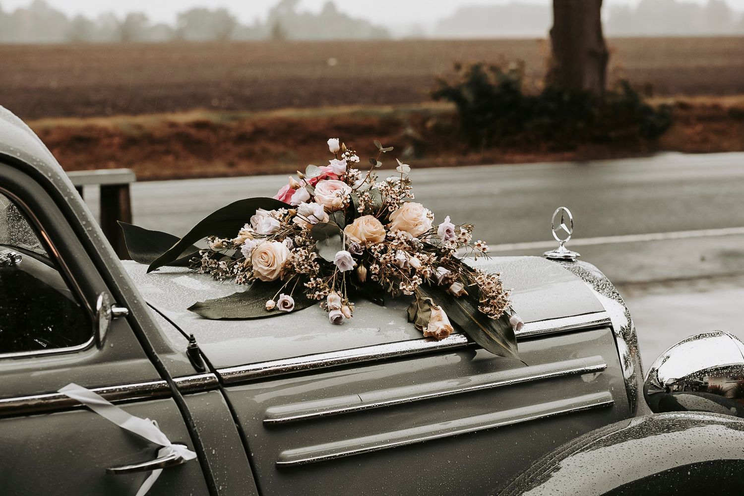 Vintage gray car hood adorned with a romantic bouquet of pink and white flowers on a rainy day.