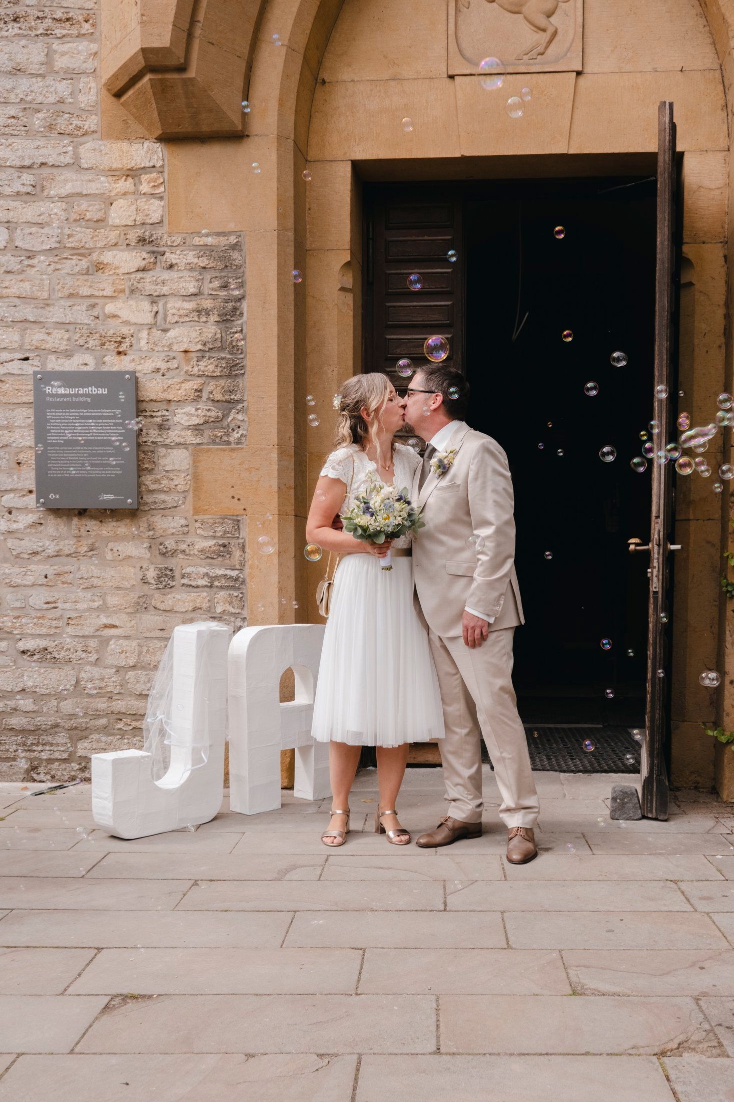 Newlyweds share a kiss outside stone building entrance with decorative white letter initials and floating bubbles.