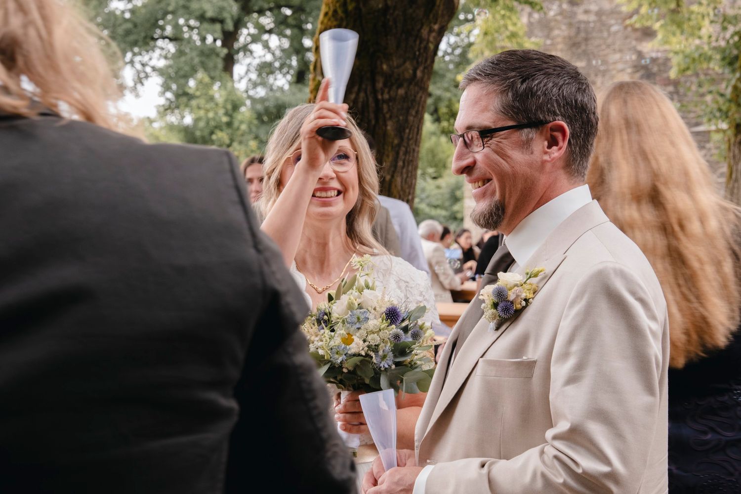 Bride laughs holding champagne flute while groom in beige suit smiles during outdoor wedding celebration.