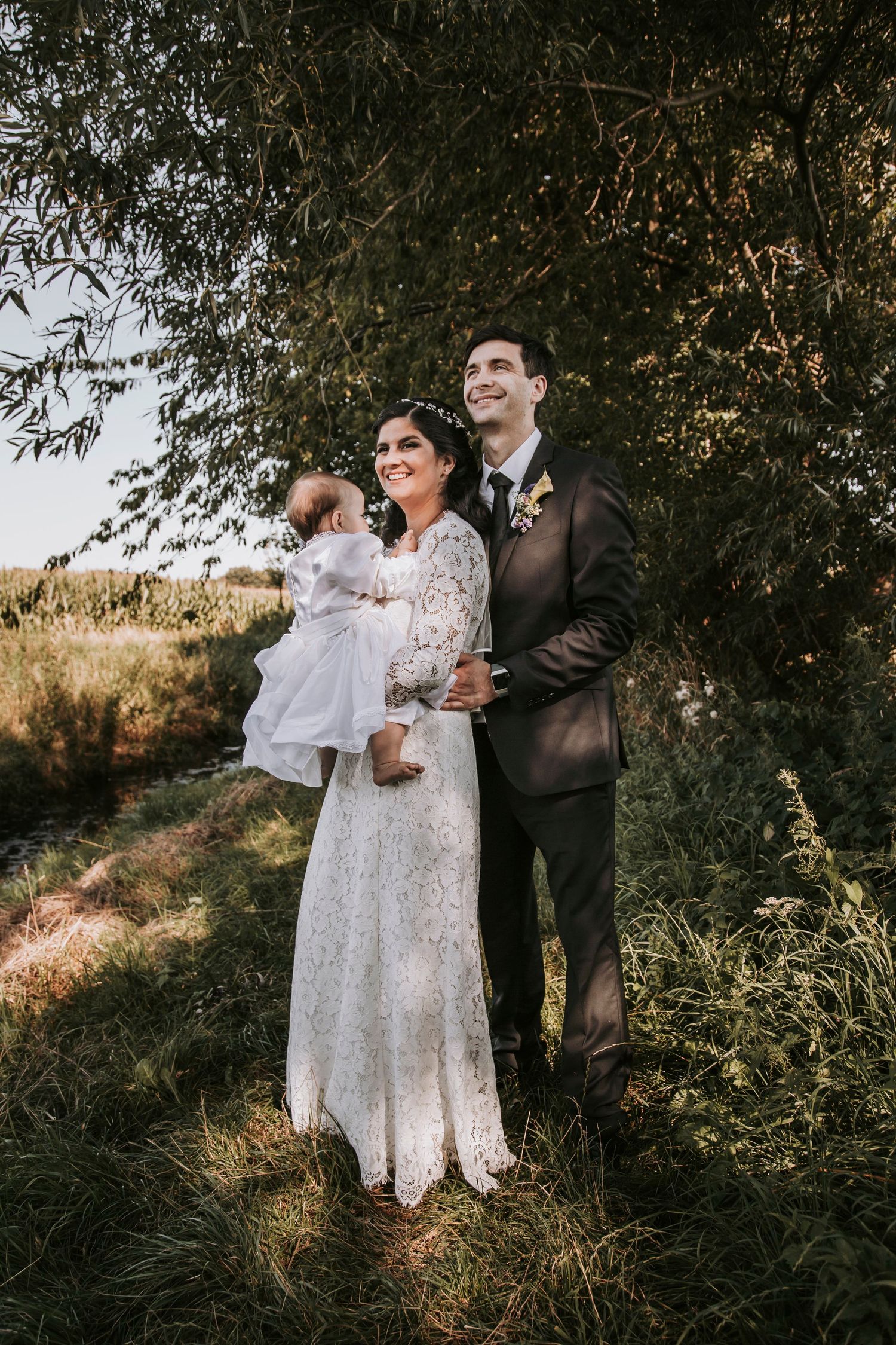 Smiling bride and groom holding infant in white dress standing under willow tree in field.