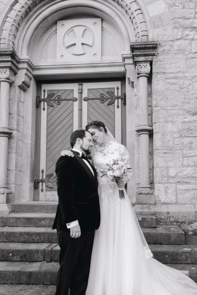 Bride and groom share a tender moment on church steps, with bride holding bouquet in classic black and white wedding photo.