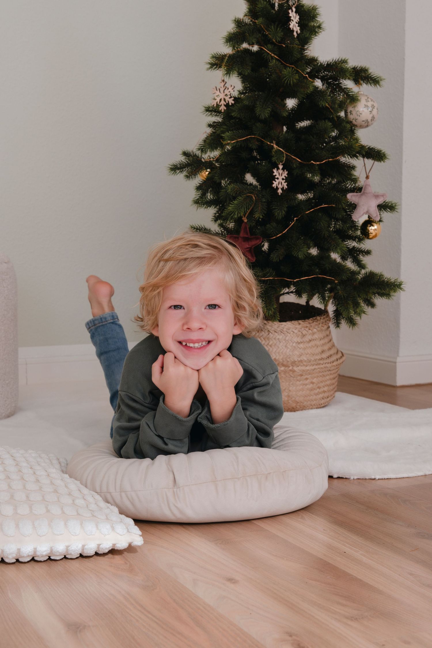 Cheerful blonde child resting on round floor cushion smiling in front of Christmas tree.