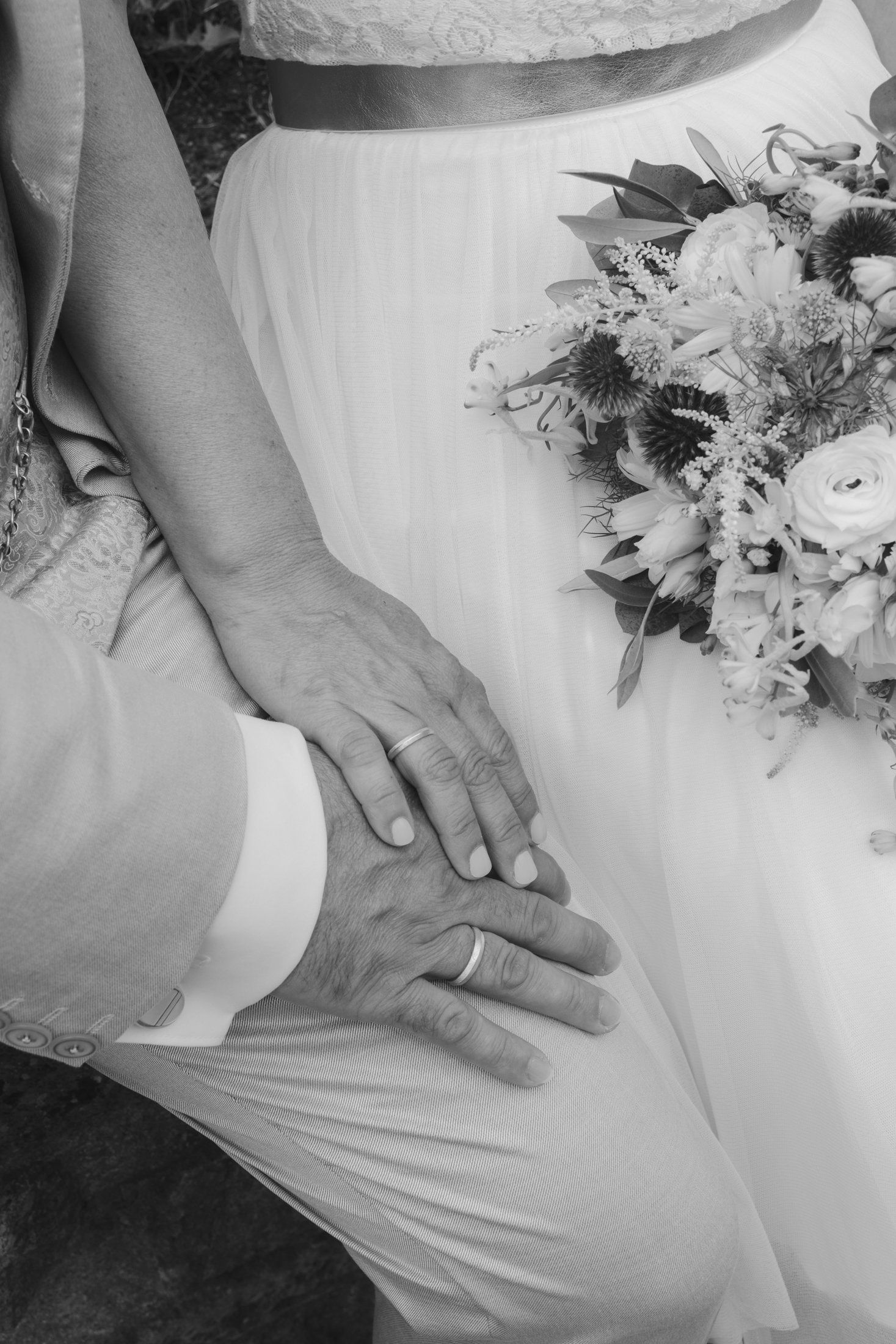 Black and white close-up of newlyweds hands with wedding rings resting on brides white dress beside bouquet.