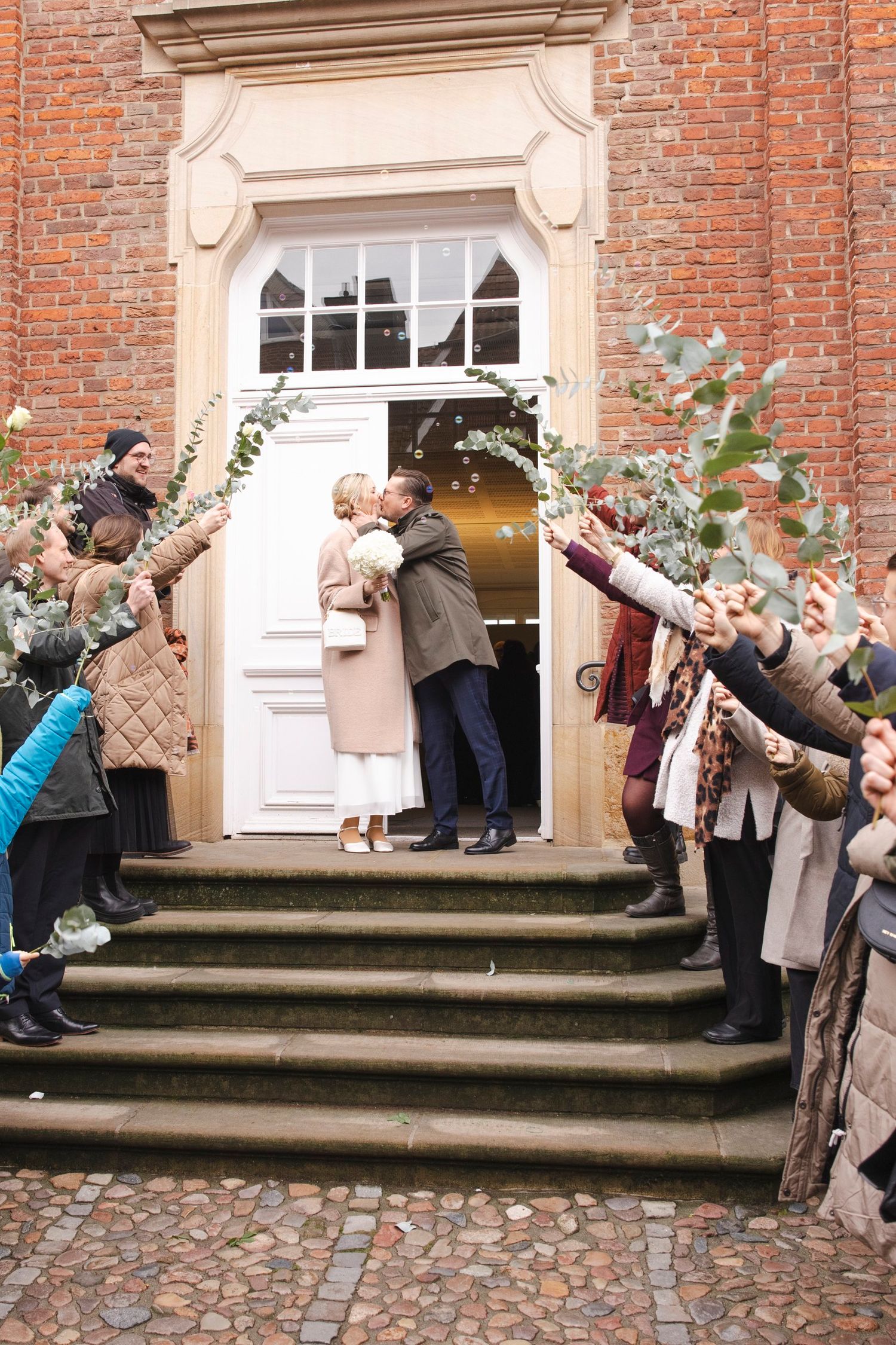 Newlyweds share a kiss on steps of brick building as guests form a floral arch.