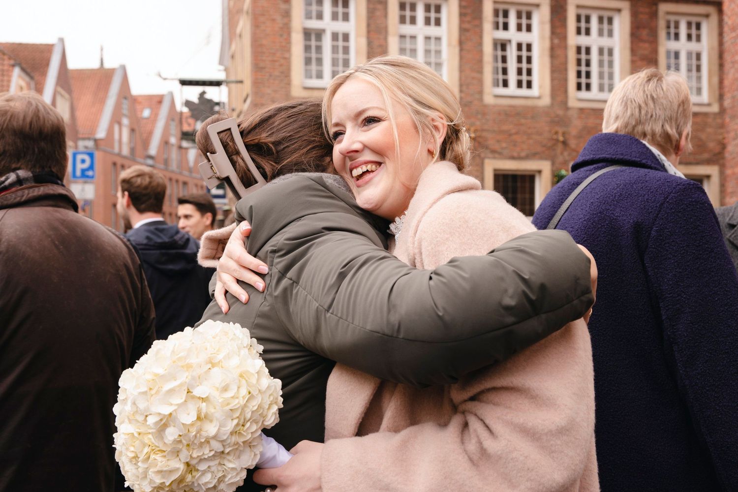 Smiling blonde bride in pink coat joyfully embraces a guest while holding white hydrangea bouquet outdoors.