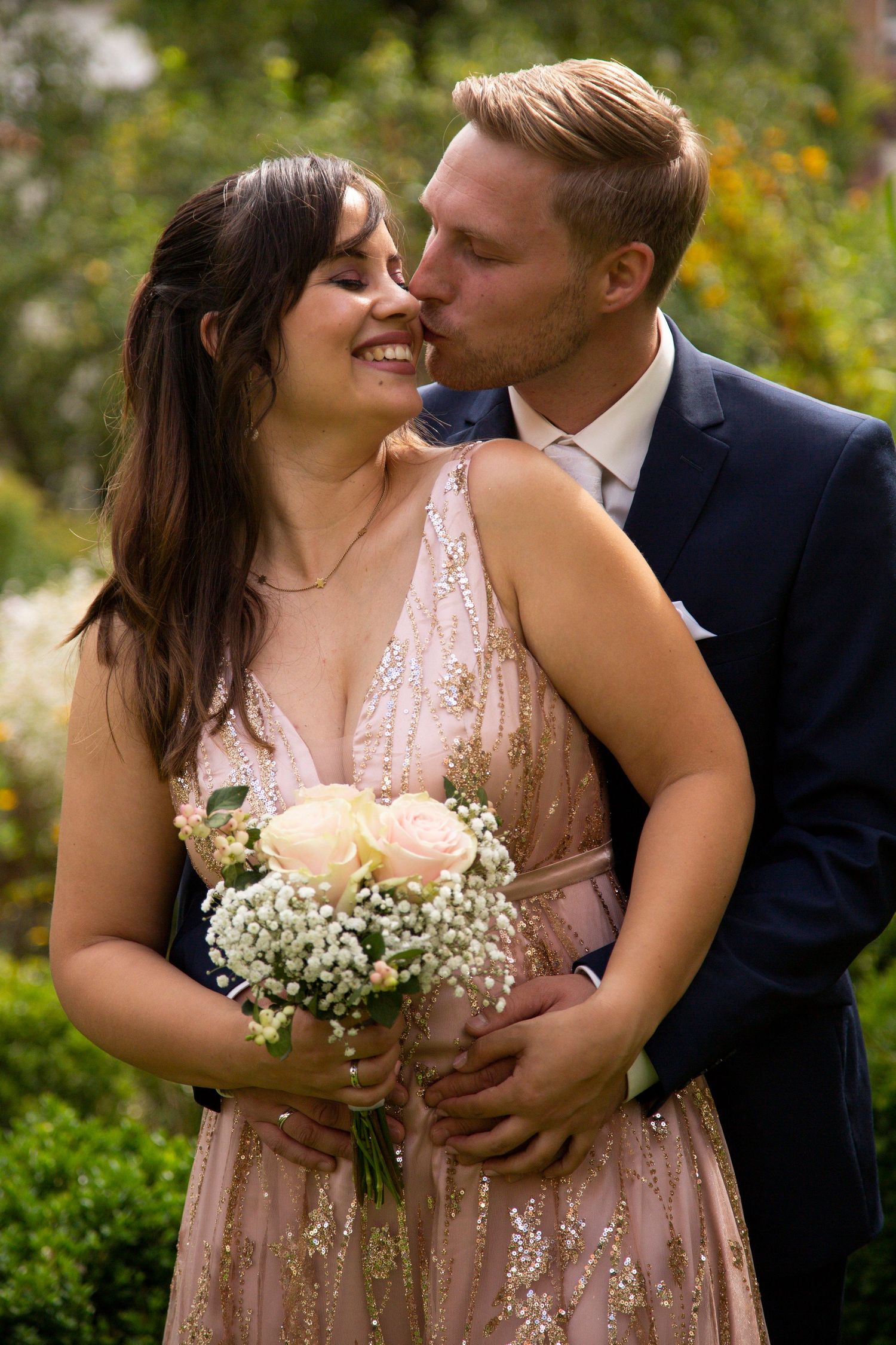 Groom in blue suit kissing smiling bride holding pink rose bouquet in garden setting.