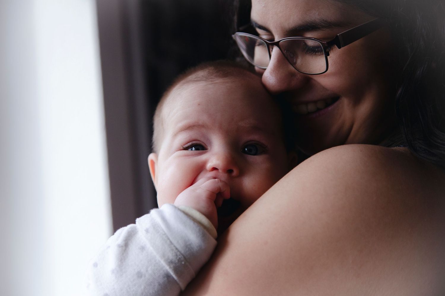 Smiling mother with glasses tenderly holding and cuddling her happy baby near a bright window.