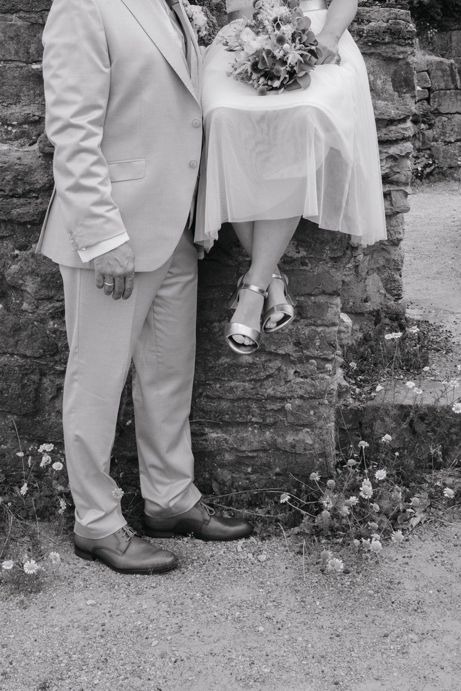 Black and white photo of bride and groom standing by stone wall, showcasing wedding attire and bridal bouquet.