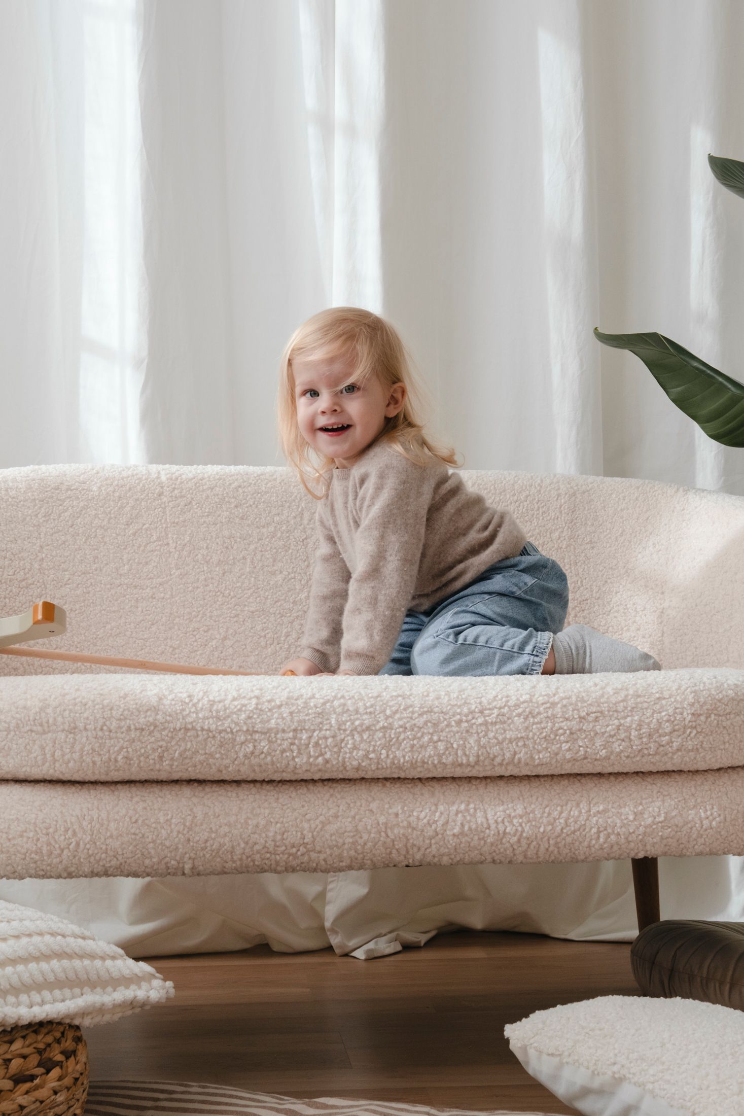 Happy toddler girl climbing on a cream boucle sofa in a bright living room.