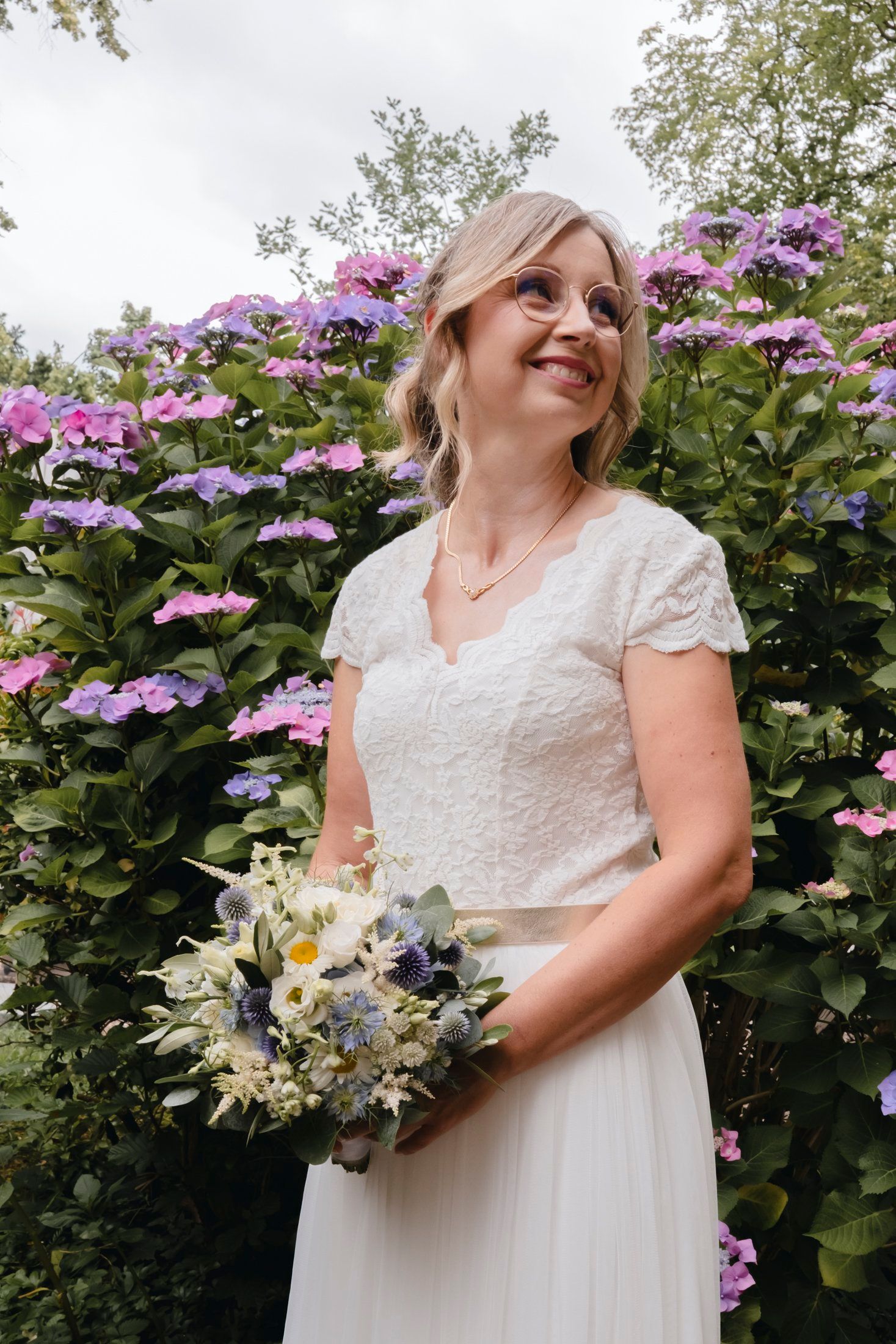 Smiling blonde bride in lace wedding dress holding bouquet, standing before colorful hydrangea bushes outdoors.