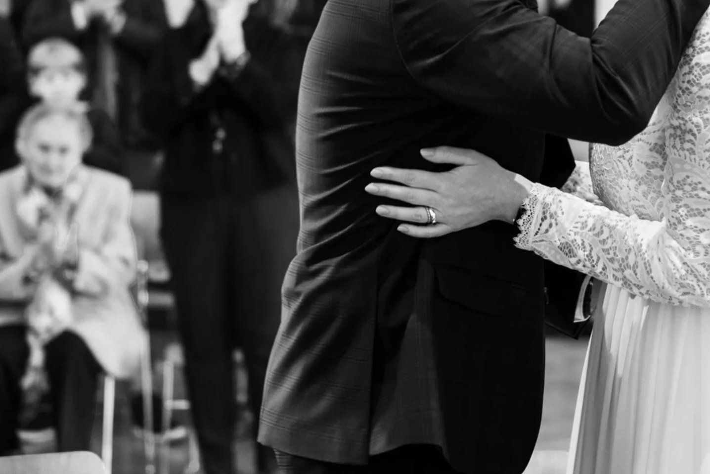 Black and white close-up of bride with wedding ring embracing groom in suit during wedding ceremony.