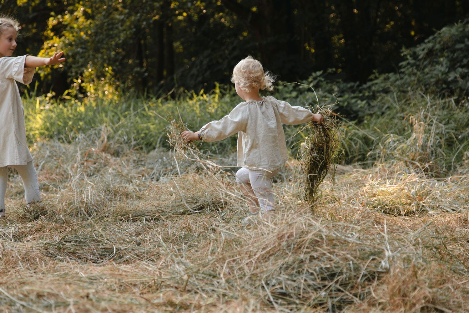 Two young children playing and running through a hay field near a lush green forest.