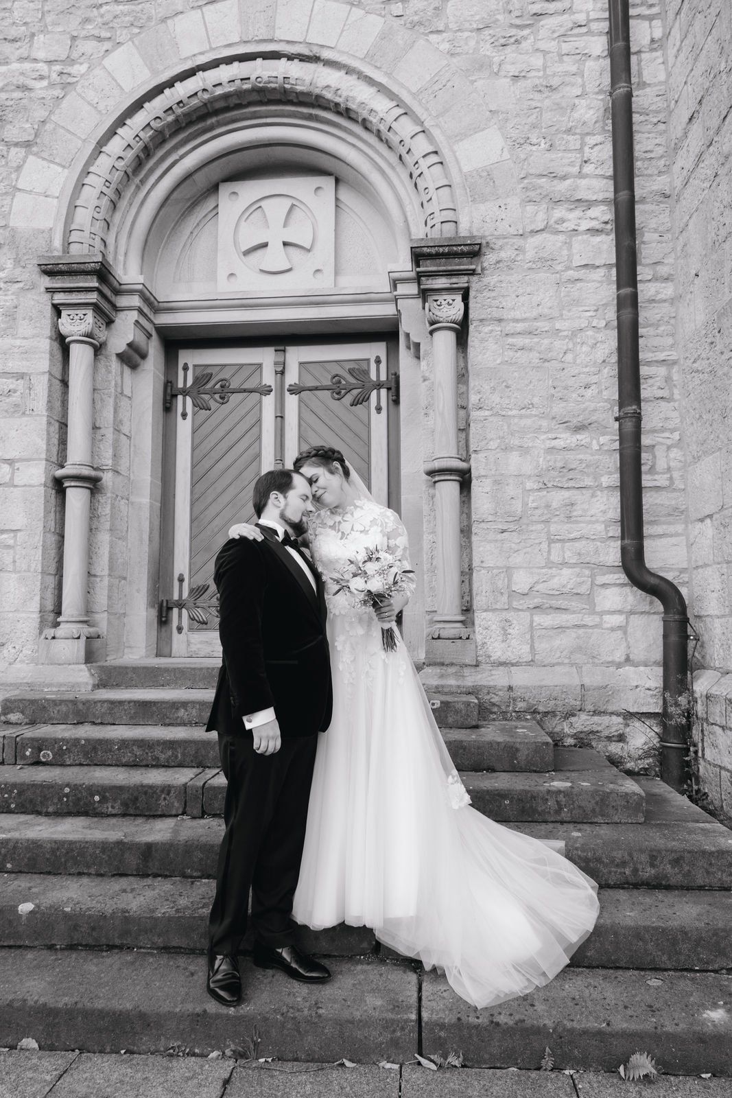 Newlyweds embrace romantically on stone church steps, bride's gown flowing in the wind in elegant black and white portrait.
