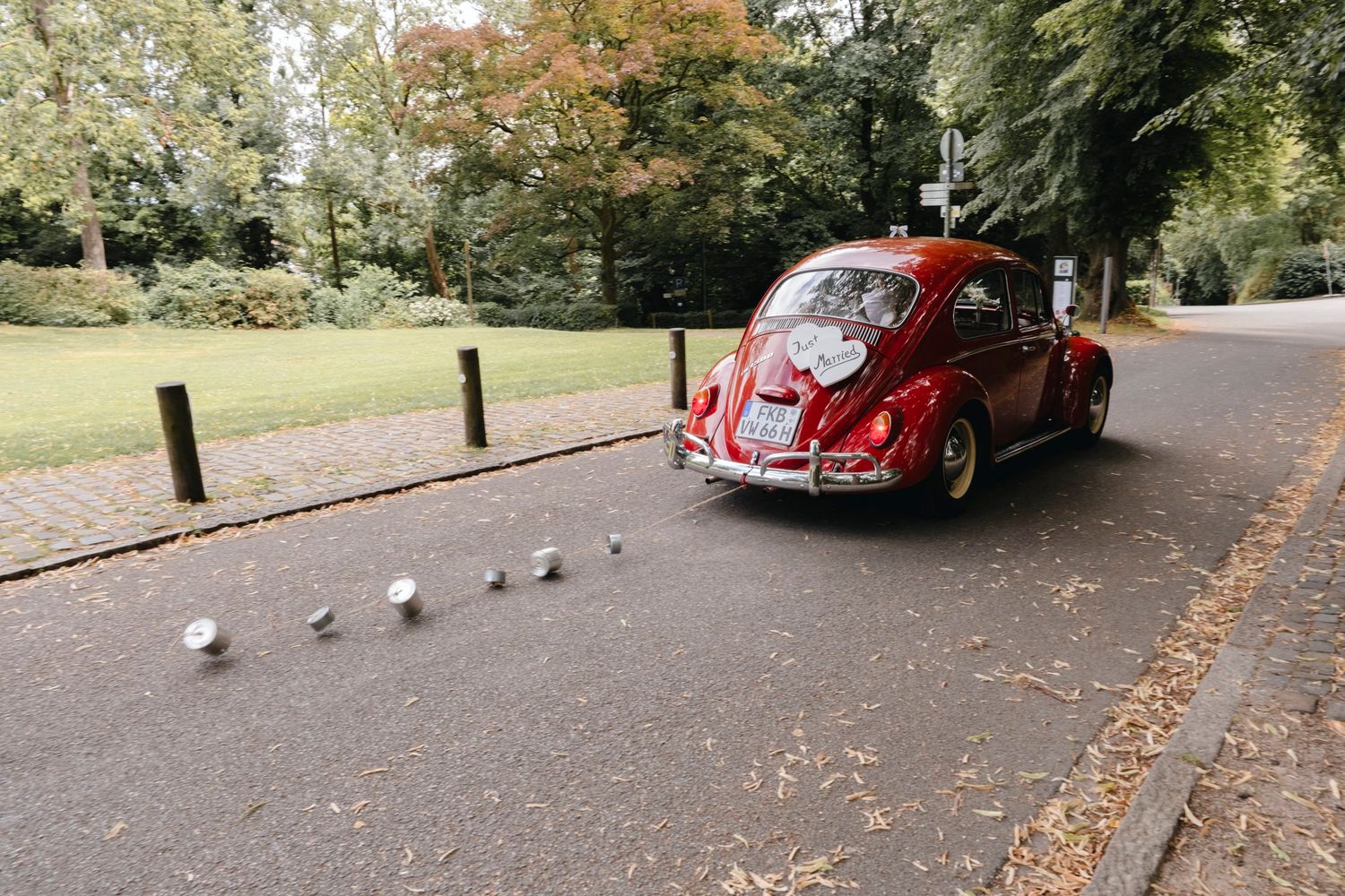 Vintage red VW Beetle wedding car driving through a tree-lined park pathway.