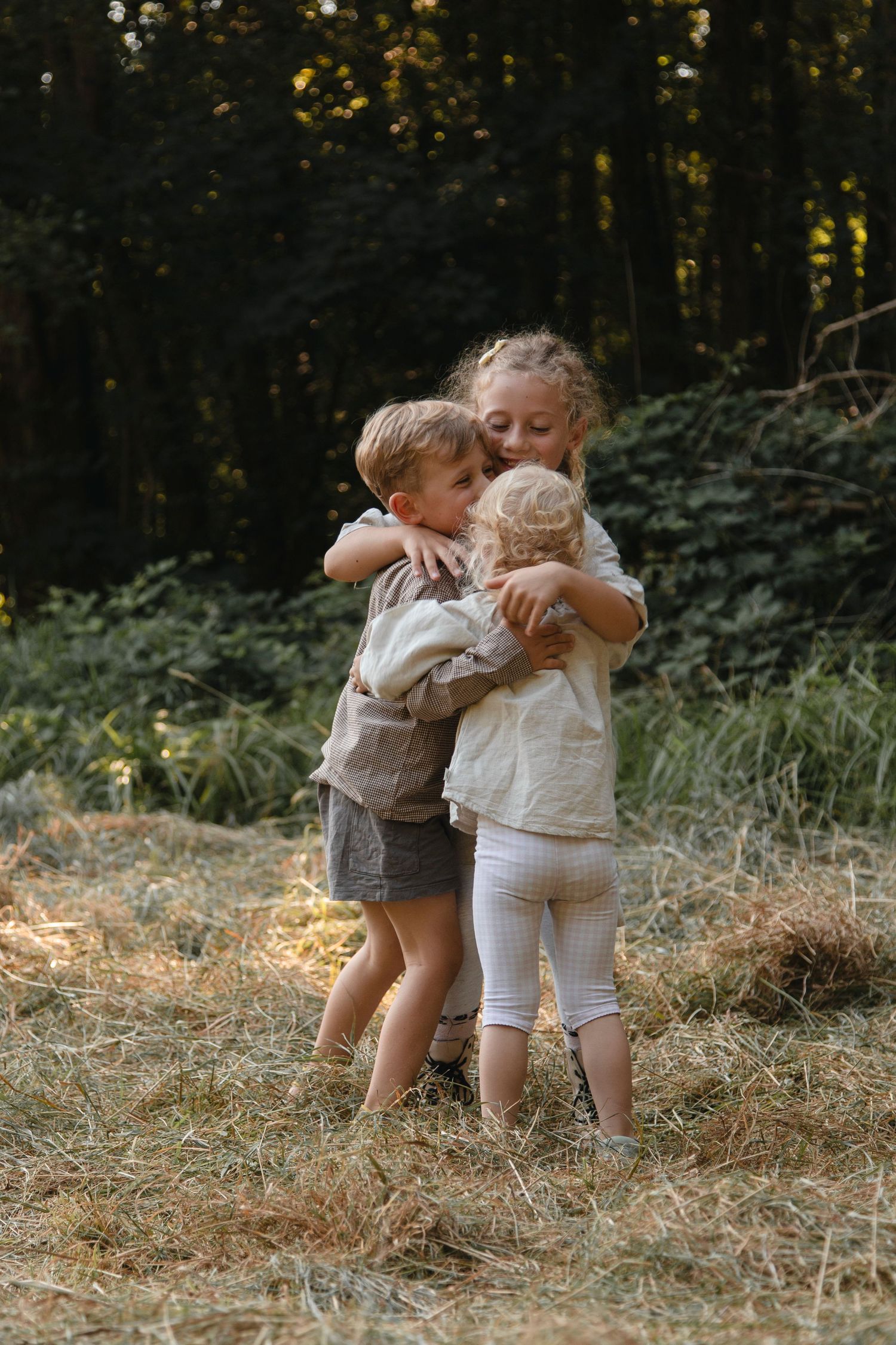 Three young siblings sharing a warm group hug in a sunlit forest clearing.