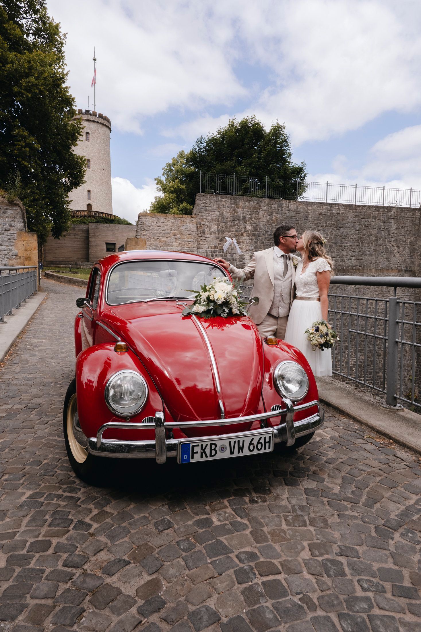 Wedding couple kissing beside a decorated vintage red VW Beetle on cobblestone street.