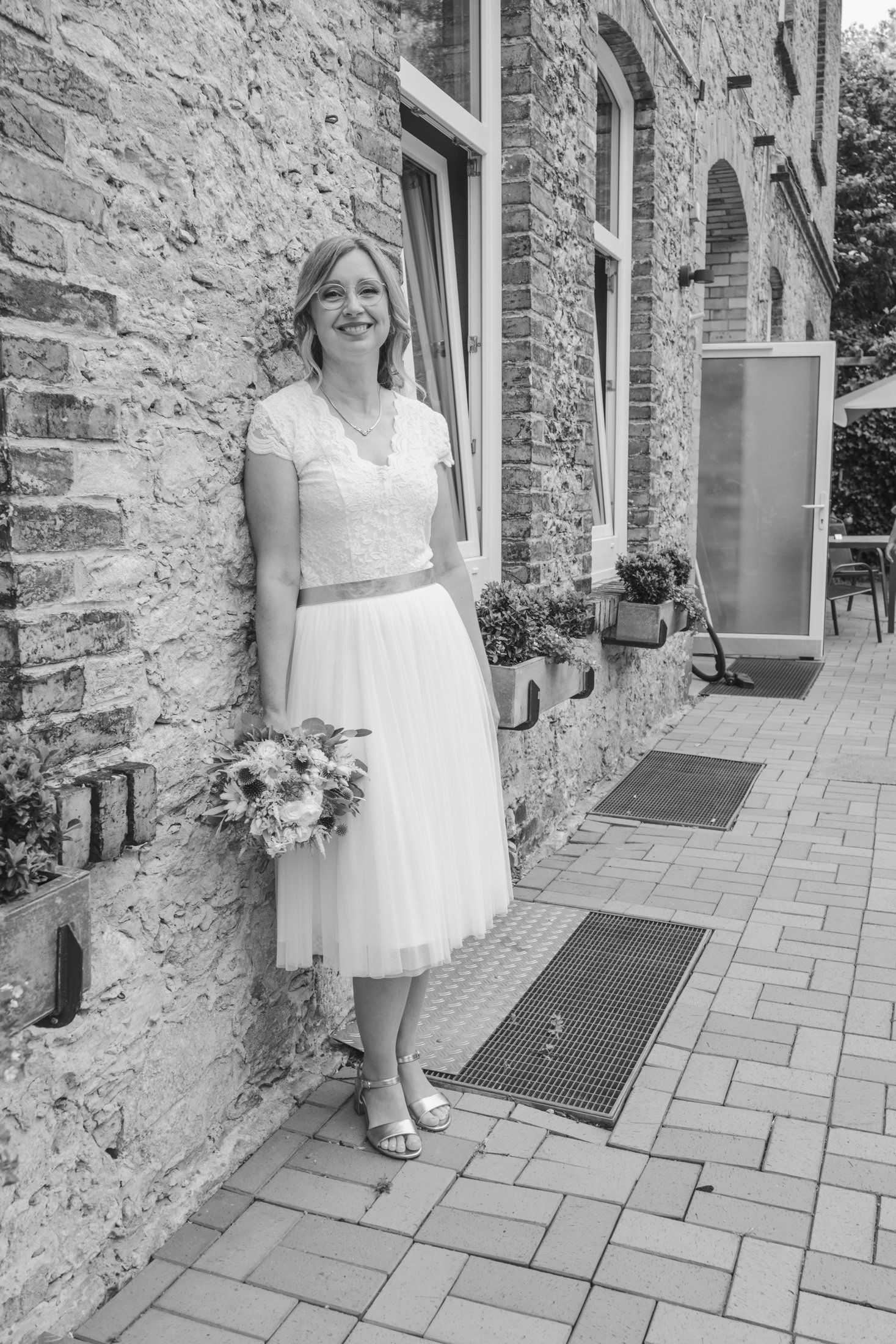 Black and white portrait of smiling bride in white dress holding bouquet, leaning against rustic stone building exterior.
