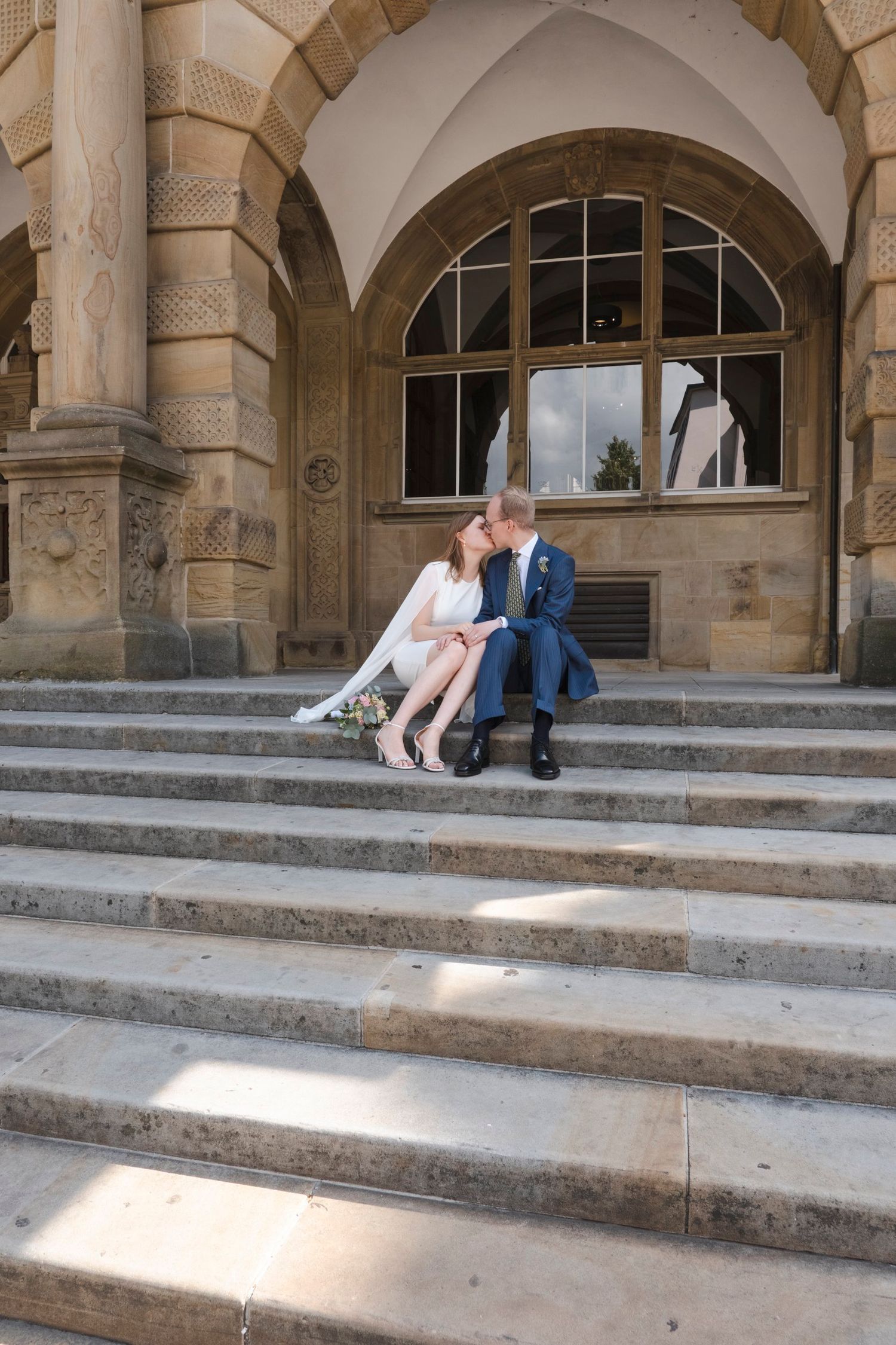 Newlywed couple sharing a kiss while seated on grand stone steps of historic building.