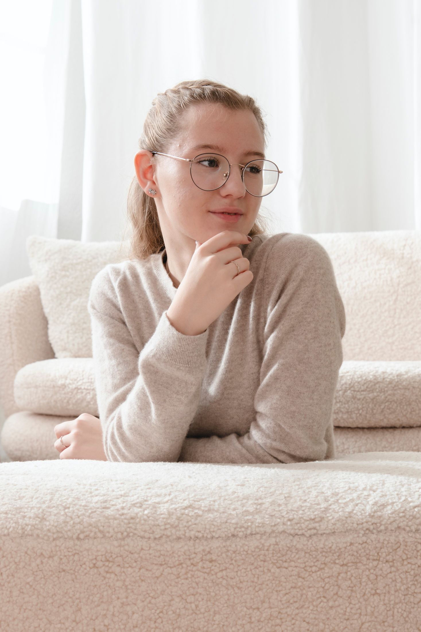 Thoughtful teenage girl with glasses resting on a cream boucle sofa in a bright room.