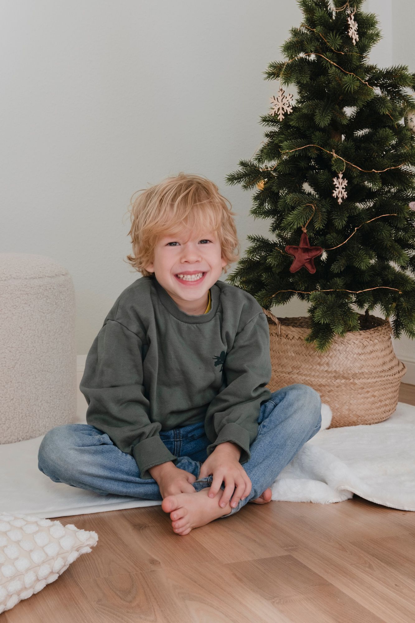 Smiling blond boy in gray sweatshirt and jeans sitting cross-legged on floor beside decorated Christmas tree.