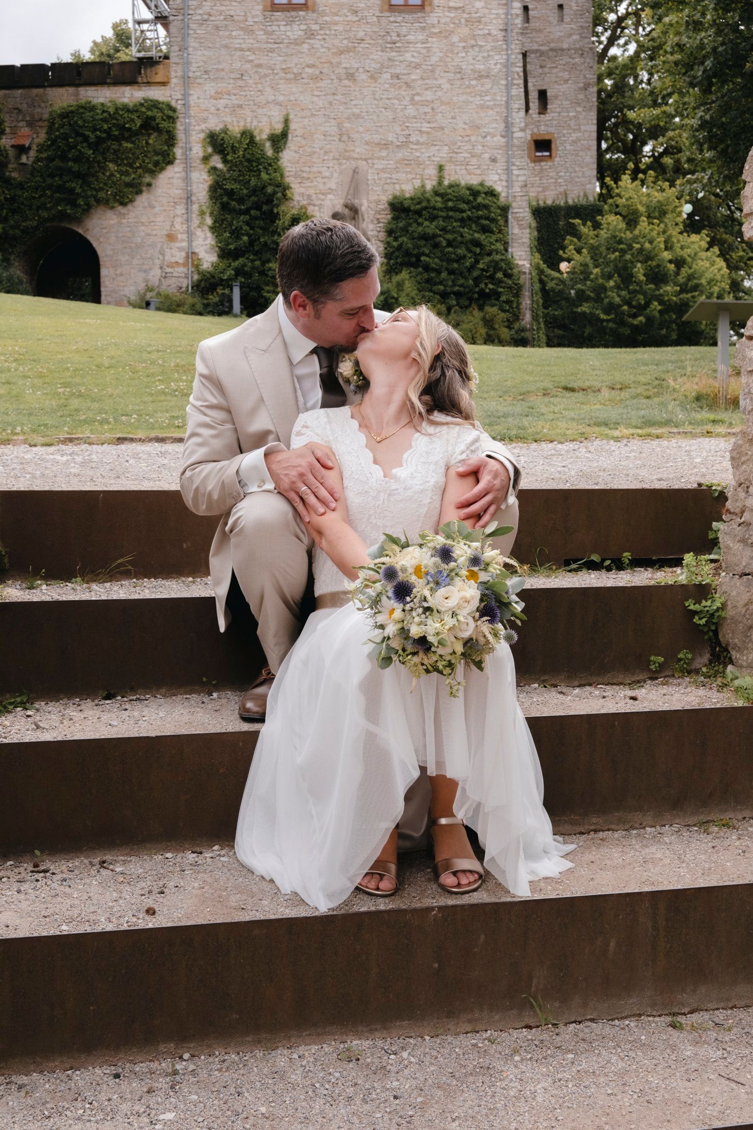 Bride and groom sharing a kiss on stone steps with castle backdrop and floral bouquet.