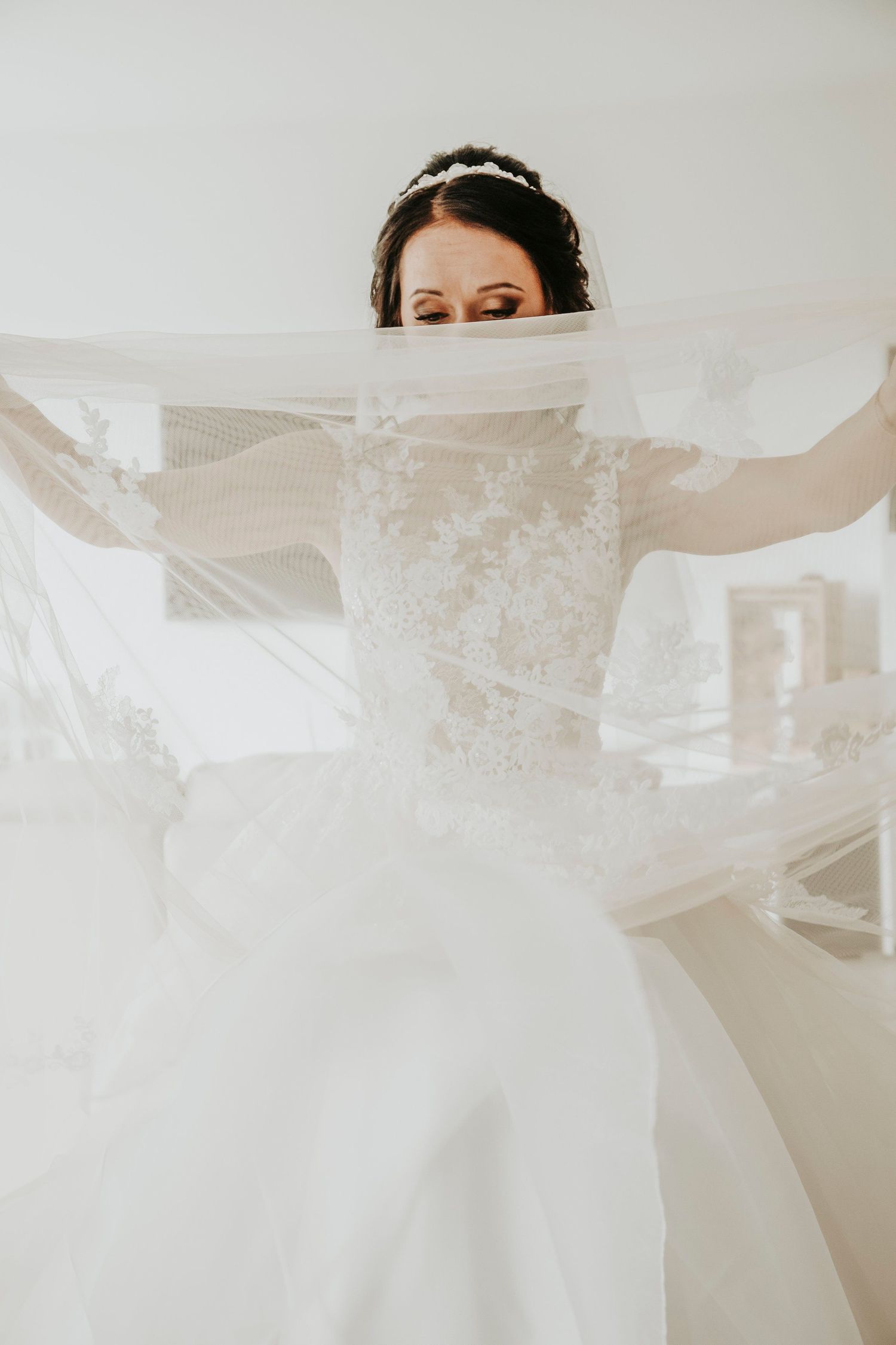Bride peeks playfully over her flowing veil with arms outstretched, wearing a lace wedding gown and floral hair piece.