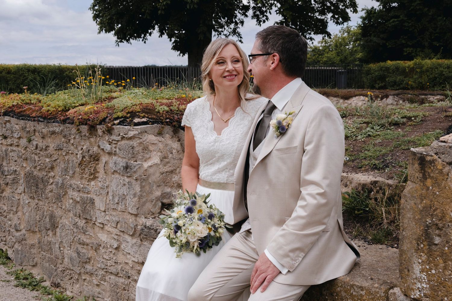 Bride and groom sit together on stone wall in garden setting, bride holding white and blue wildflower bouquet.