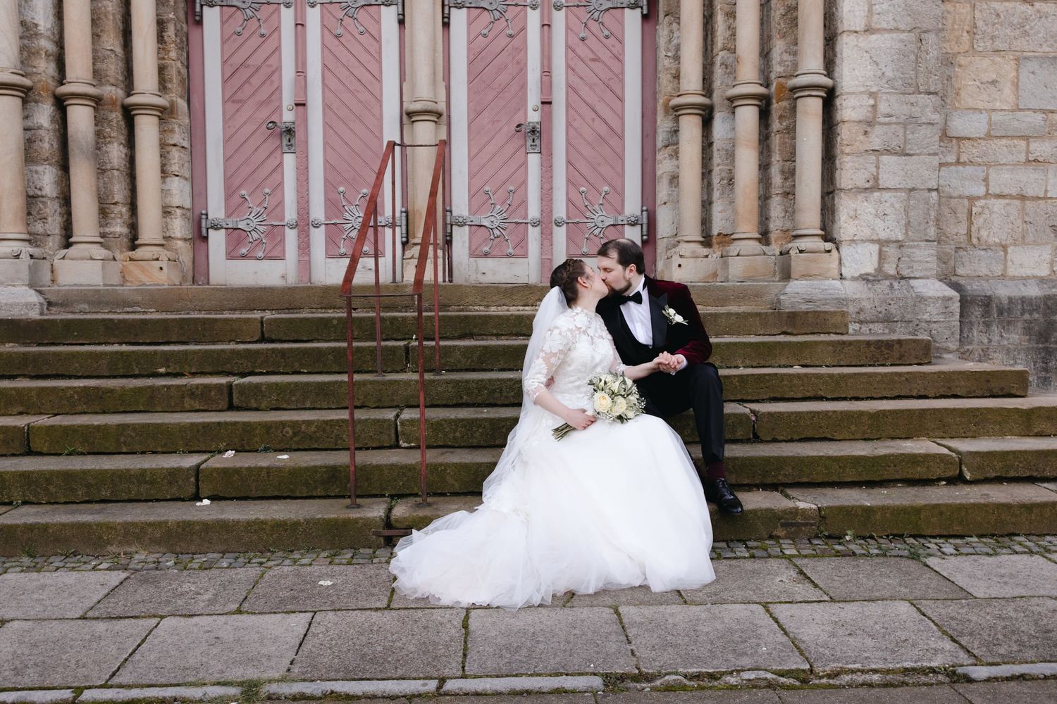 Bride and groom share a kiss while seated on church steps, bride in full tulle gown with veil in color photo.